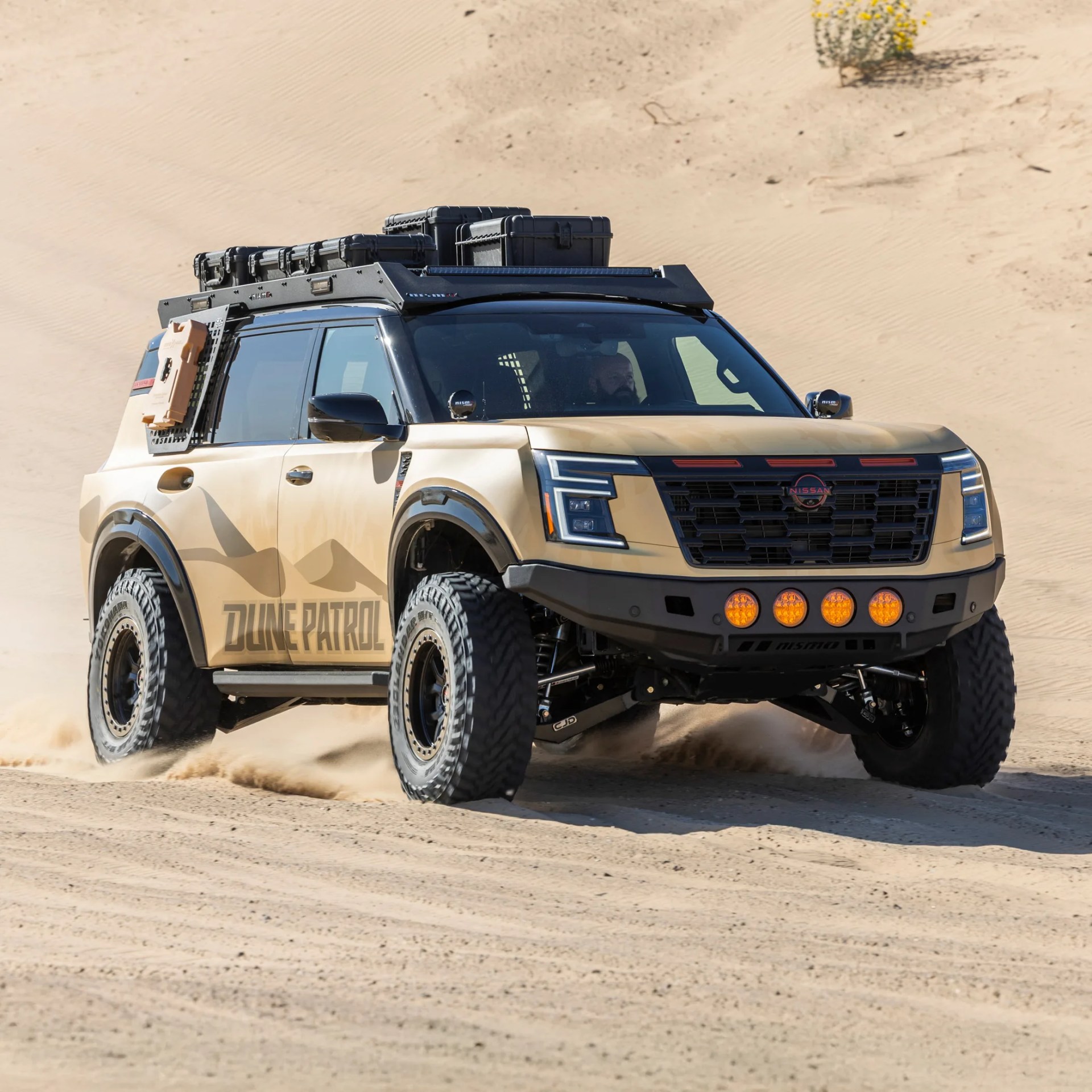 Tan Nissan SUV with "Dune Patrol" decal, off-road tires, roof rack with storage boxes, and additional front lights driving on sand dunes.