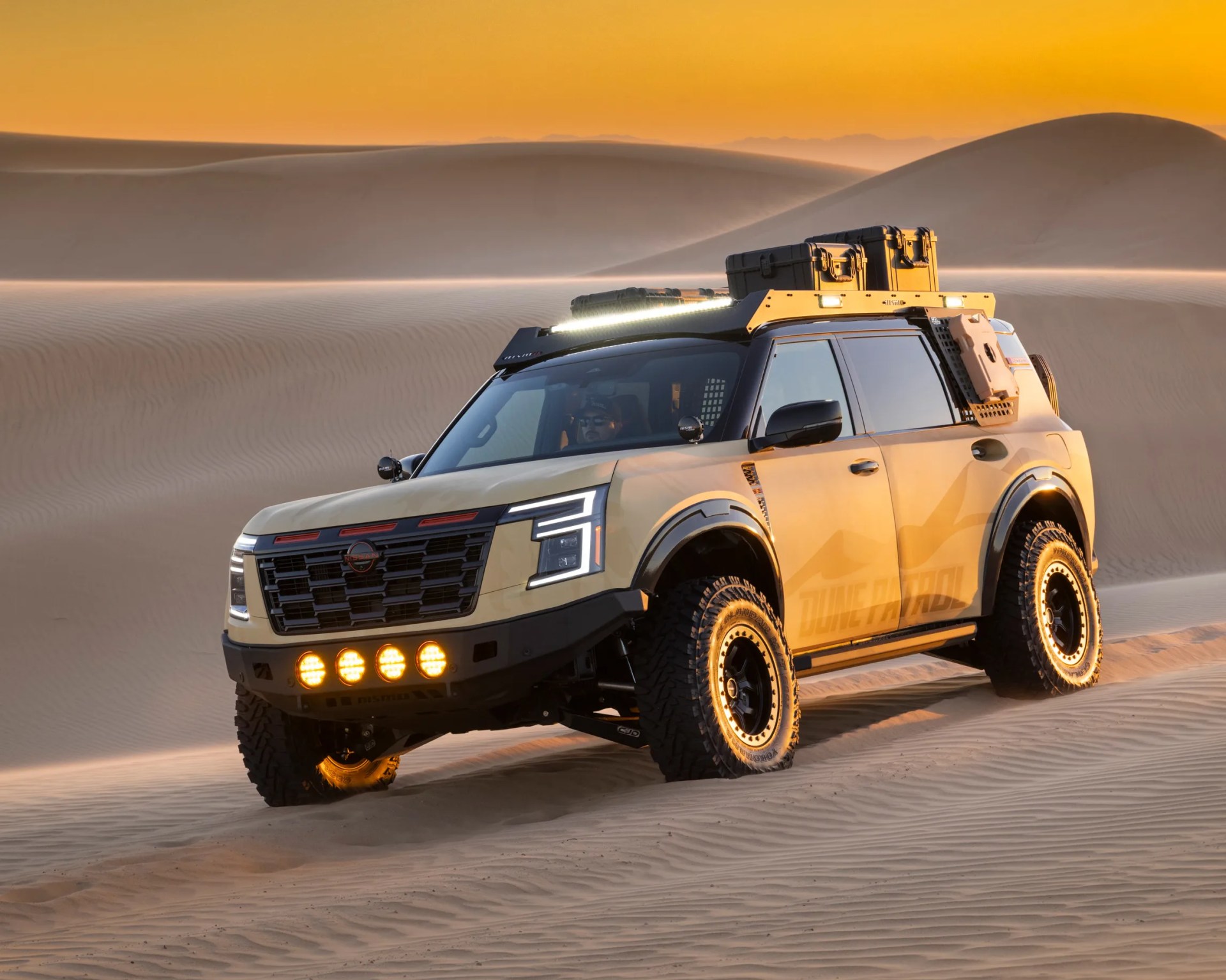 Beige Nissan SUV with off-road tires, roof rack, and additional lights driving on sand dunes at sunset.