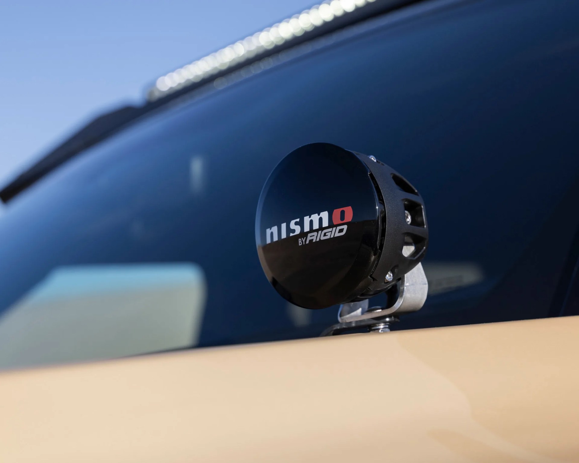 Black round Nismo by Rigid off-road light mounted on a beige vehicle with a dark windshield background.