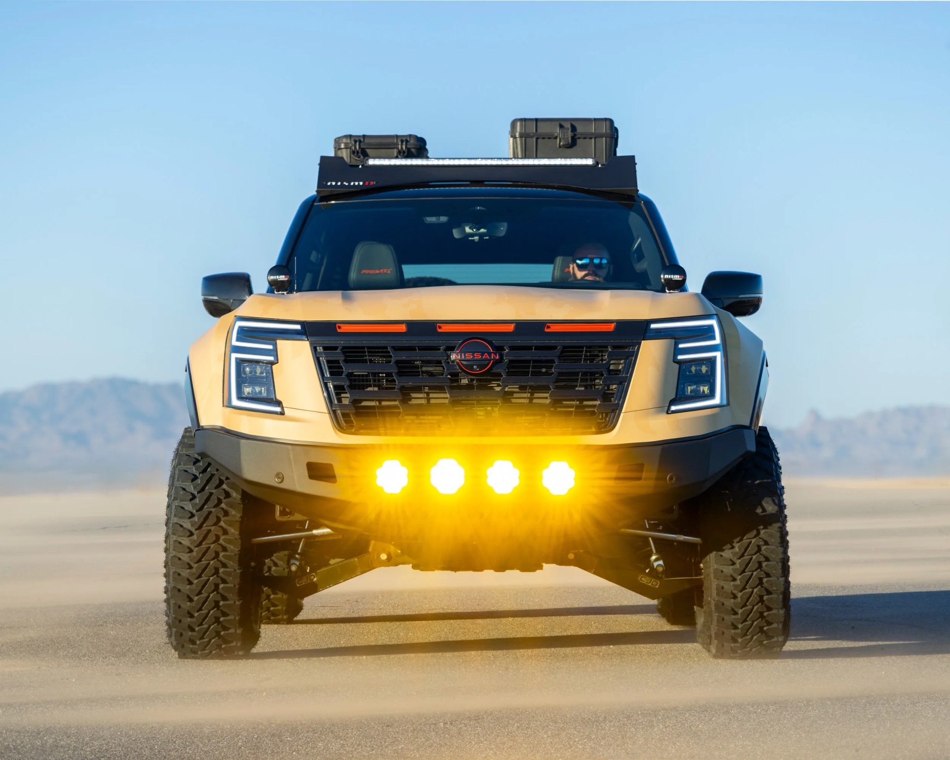 Front view of a beige Nissan off-road truck with large tires, roof storage boxes, and bright yellow auxiliary lights on a desert road.