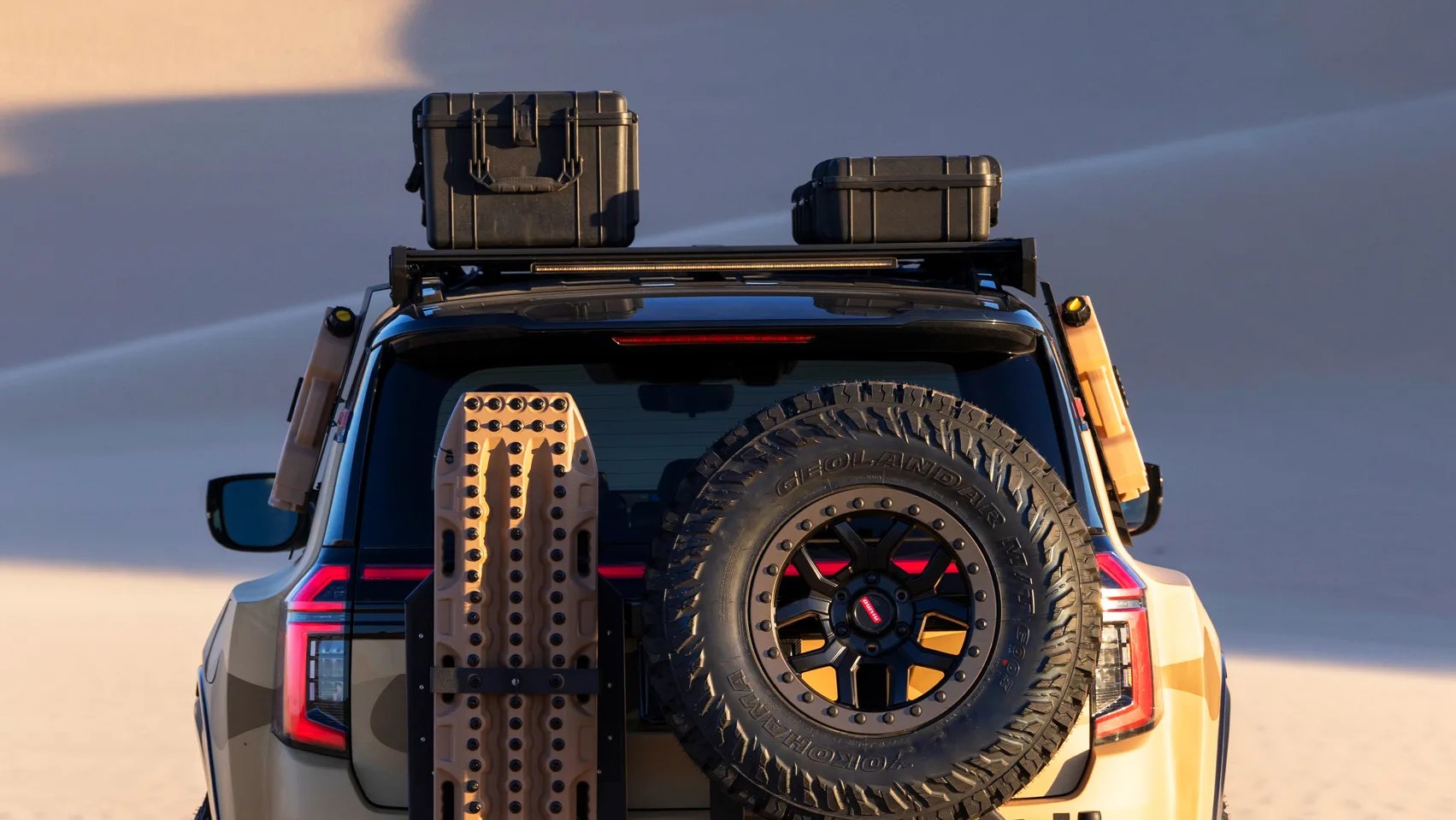 Rear view of a tan off-road vehicle with a spare Yokohama Geolandar tire, recovery boards, and two black storage cases on the roof rack.