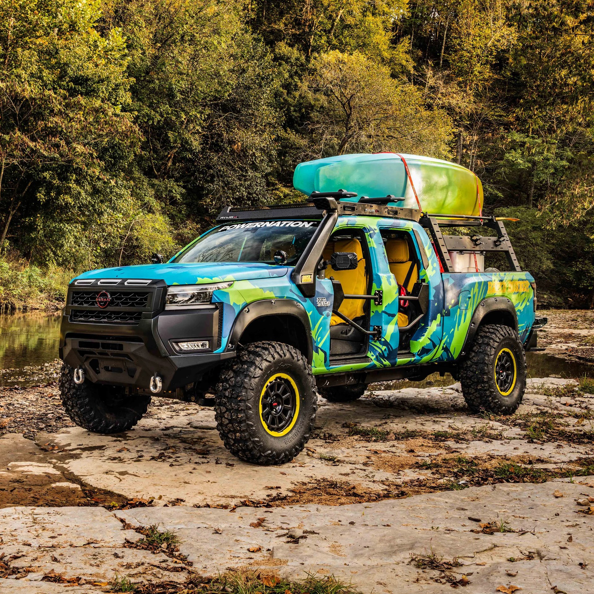 Blue and green camo Nissan pickup truck with off-road tires and two kayaks on the roof rack.