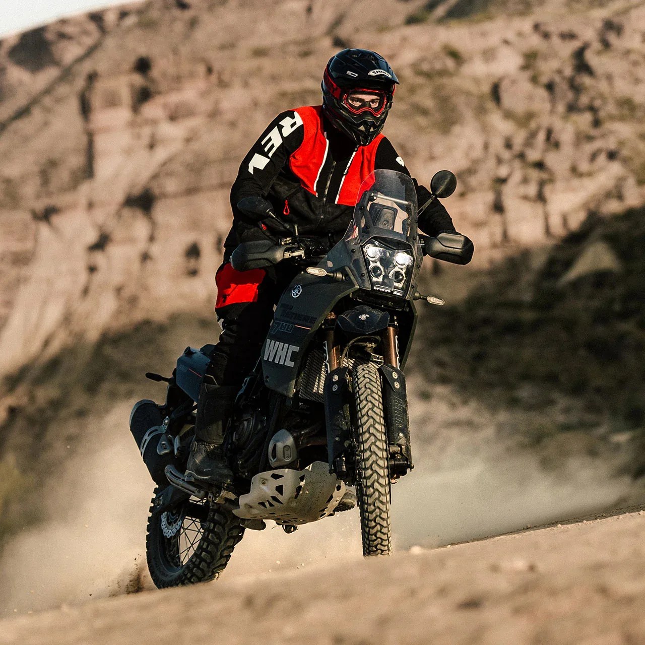 Rider in black and red gear on a black adventure motorcycle riding on a dusty dirt trail with rocky hills in the background.