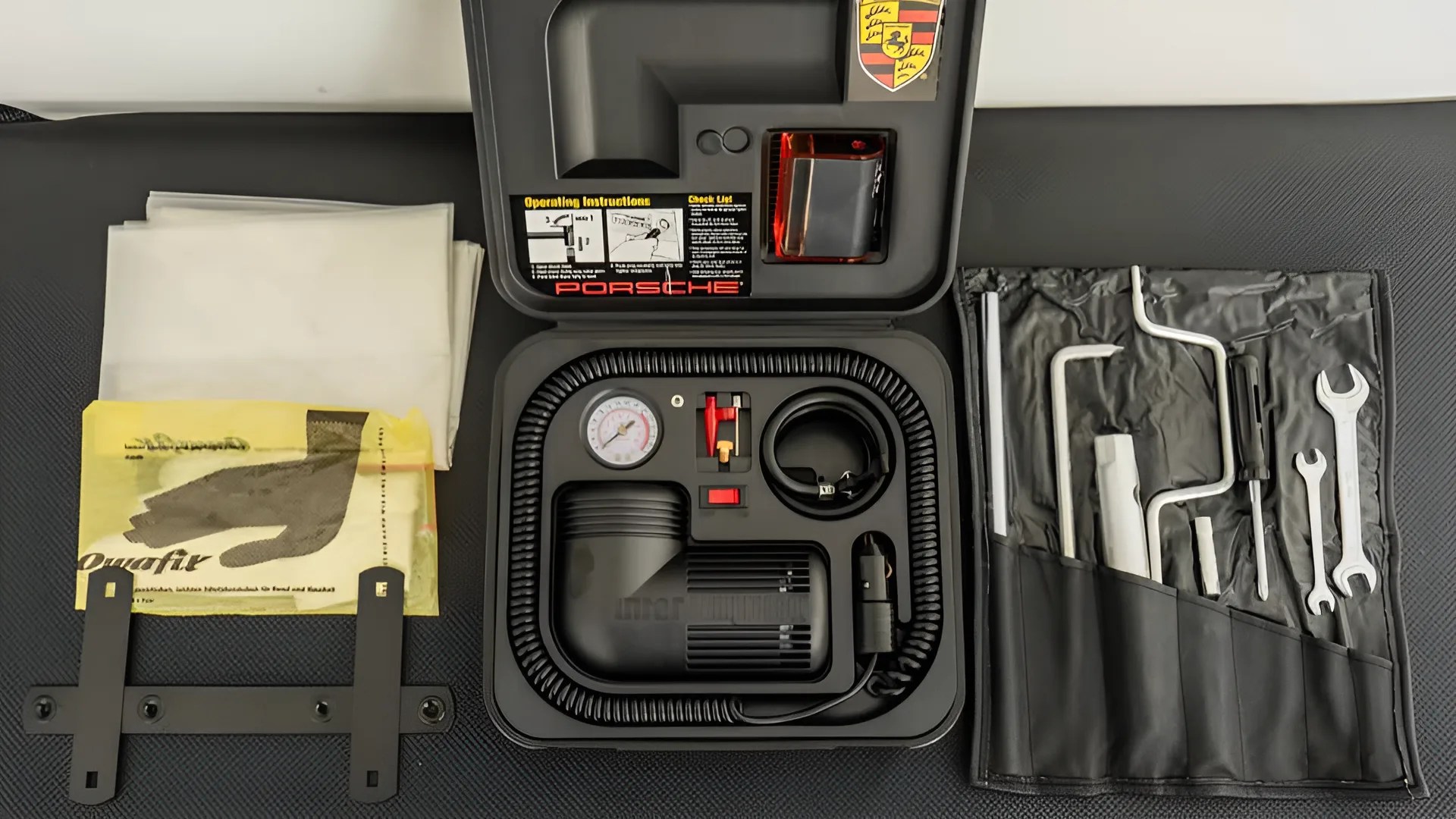 Porsche tire repair kit with air compressor, pressure gauge, tools in black case, and repair patches on a textured surface.