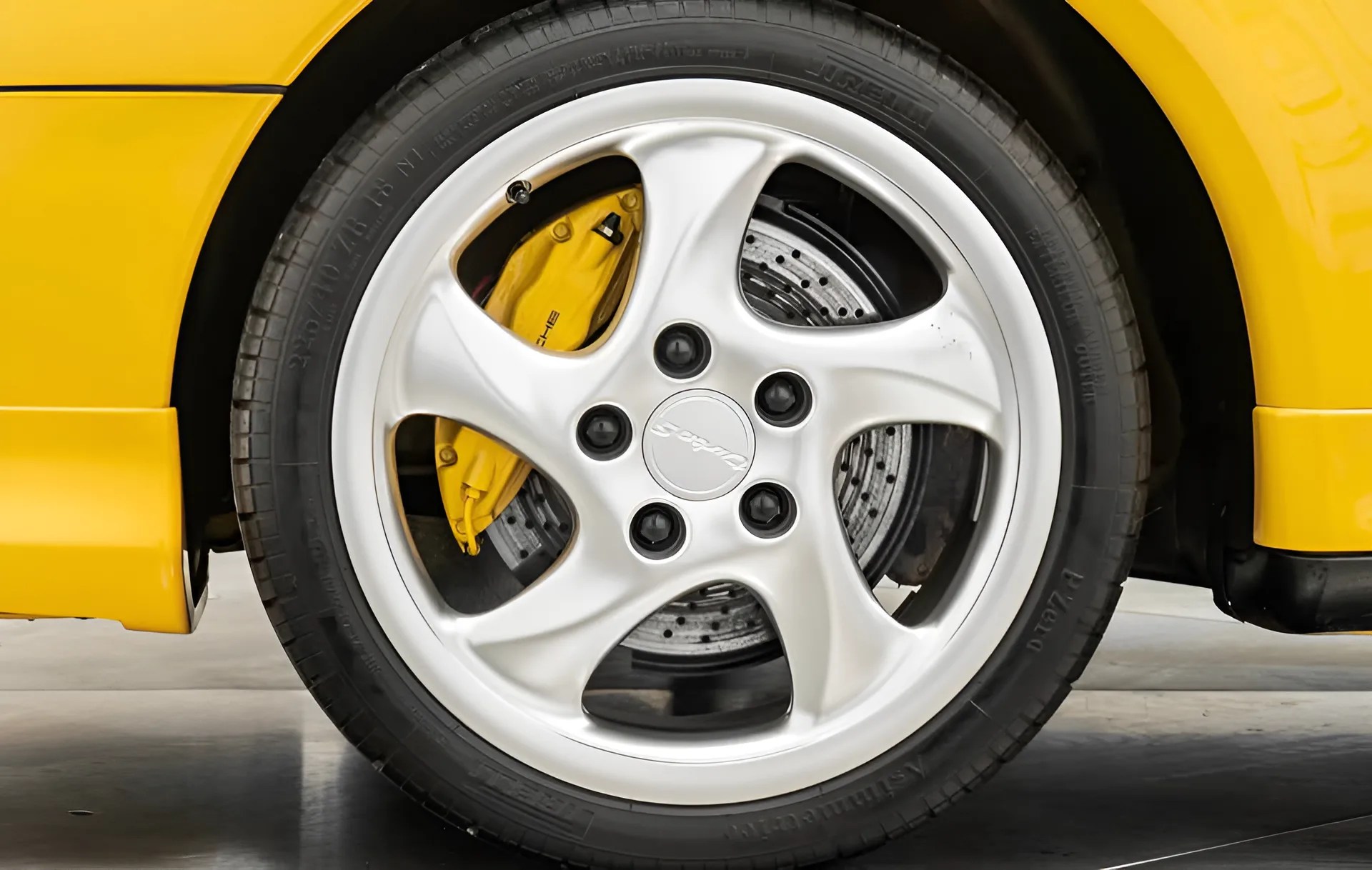 Silver five-spoke Porsche wheel with yellow brake caliper and drilled brake disc on a yellow car.