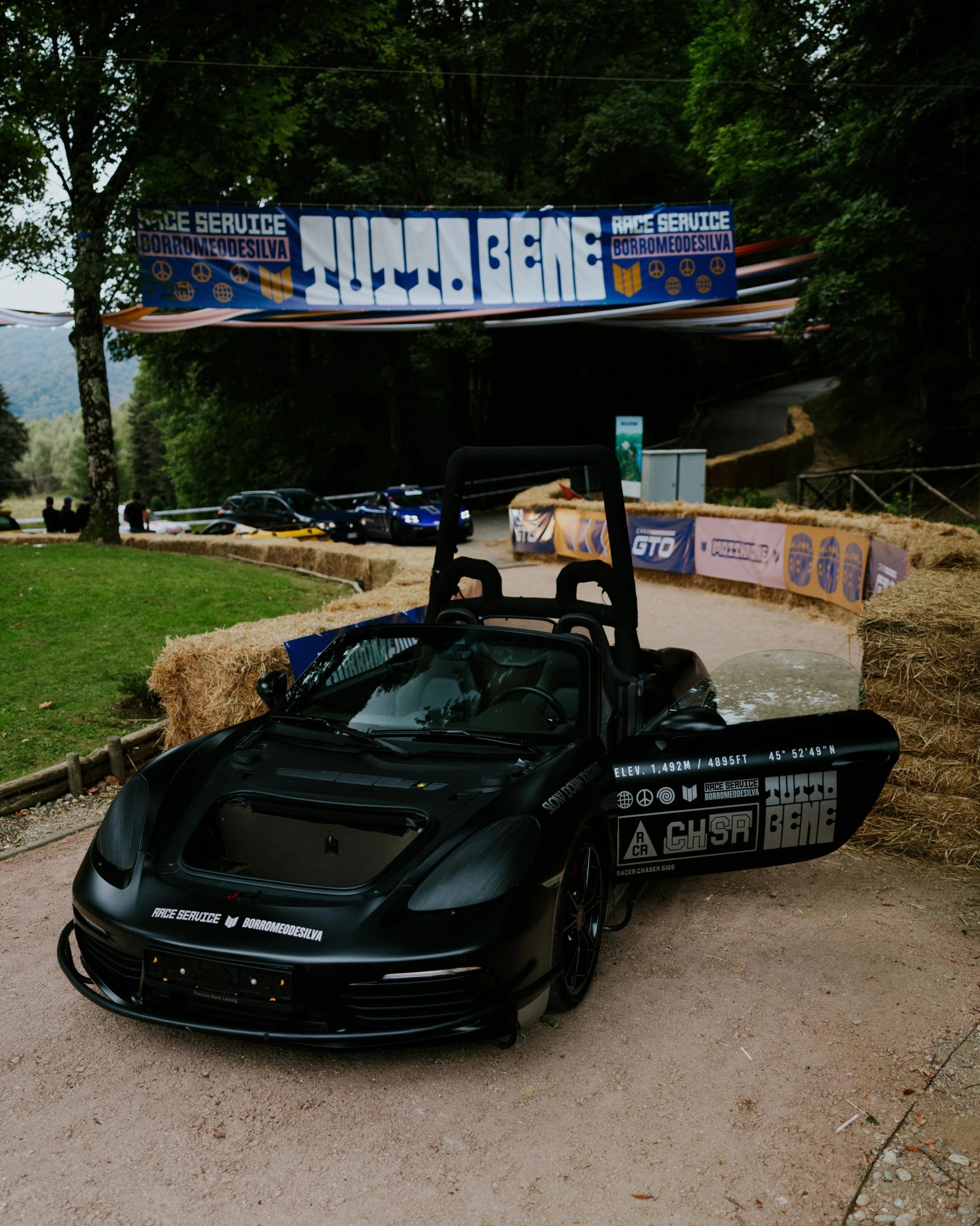 Matte black race car with open driver's door and white sponsor decals parked on a dirt track surrounded by hay bales.
