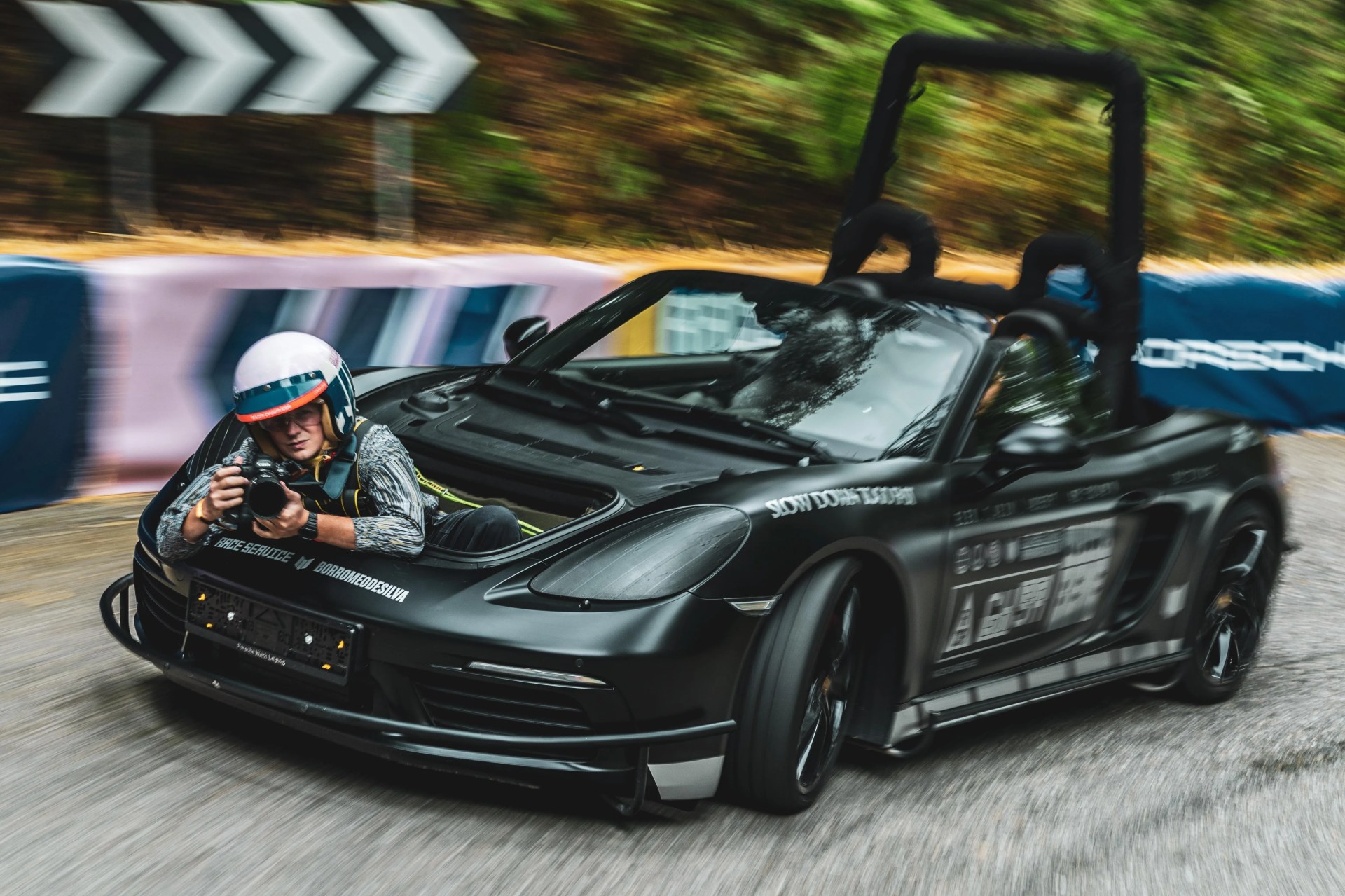 Matte black Porsche convertible with a person wearing a helmet leaning out the front holding a camera.
