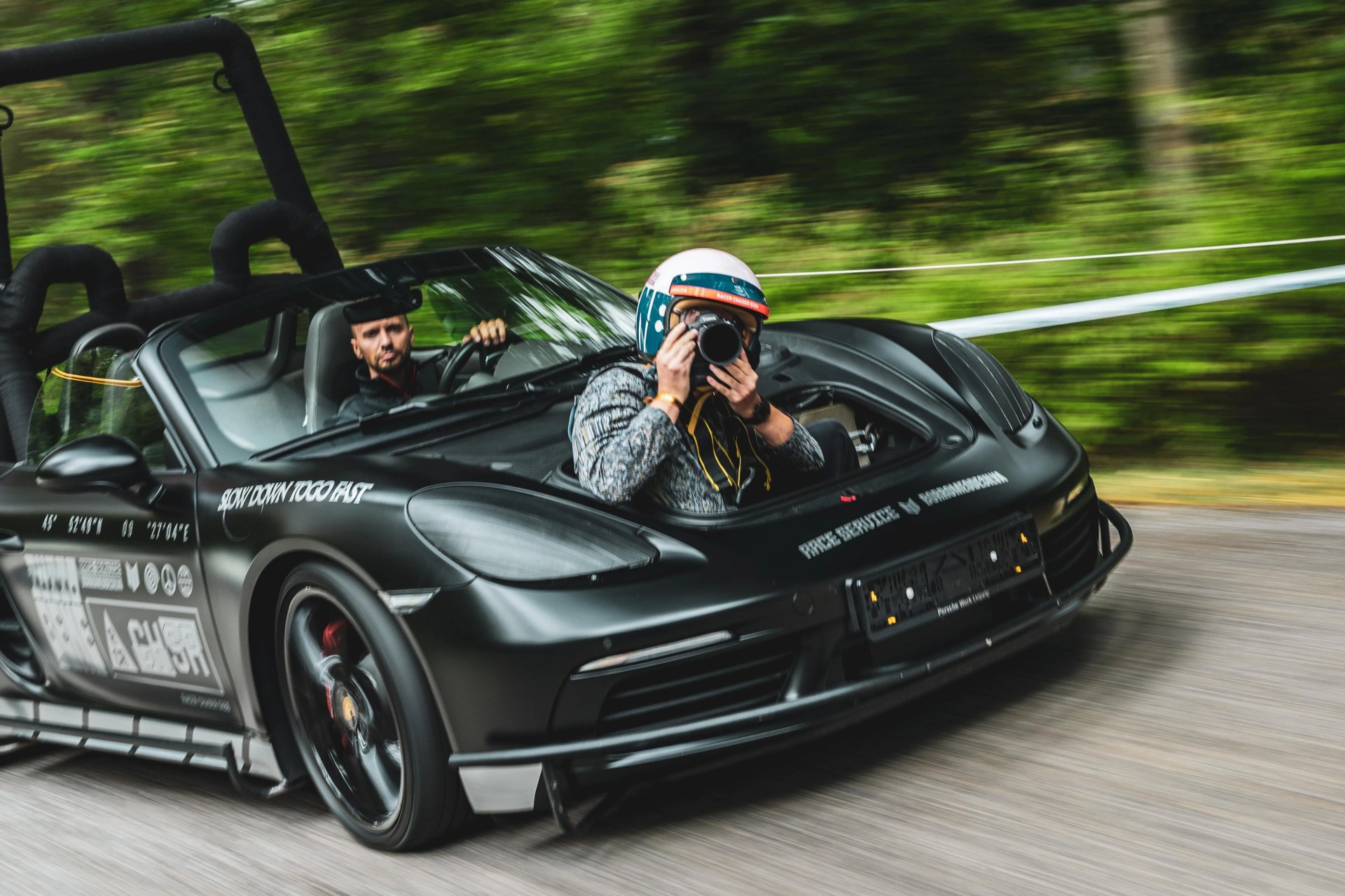Black Porsche convertible with racing decals and a person wearing a helmet holding a camera in the passenger seat.