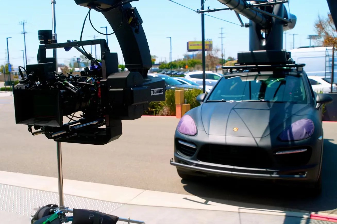 Black professional film camera mounted on a rig next to a dark gray Porsche SUV with covered headlights.