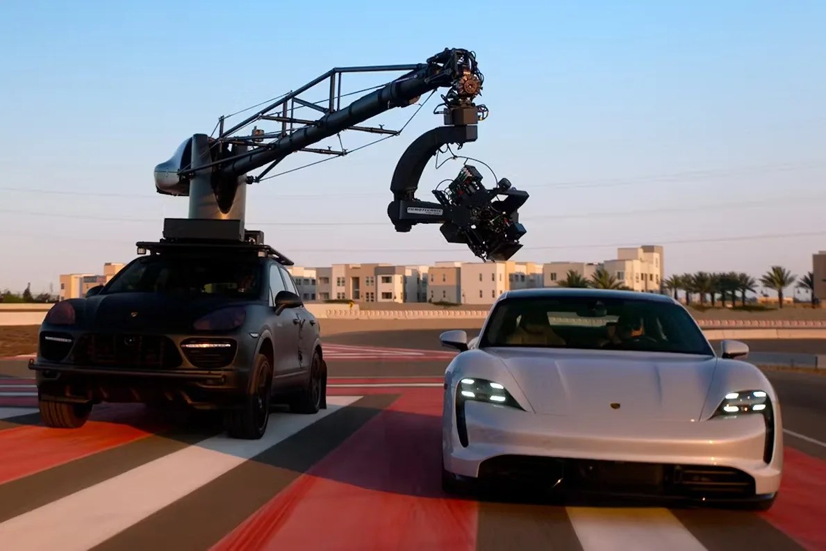 Black SUV with mounted camera crane next to a white Porsche Taycan on a racetrack.