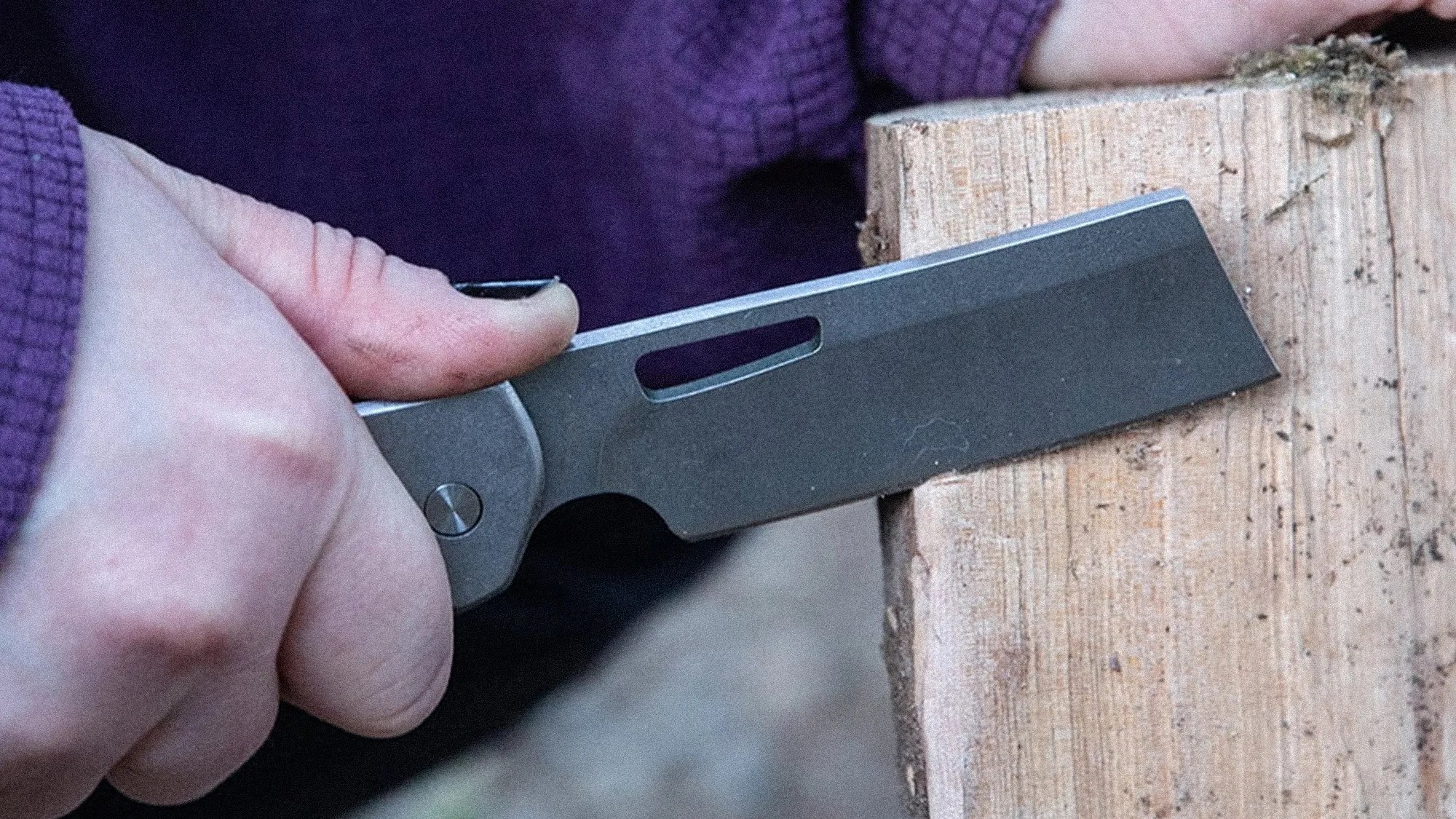 Hand holding a gray folding knife with a rectangular blade cutting into a piece of wood.