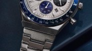 Close-up of a silver metal wristwatch with a blue bezel and white dial showing chronograph subdials and a date window.