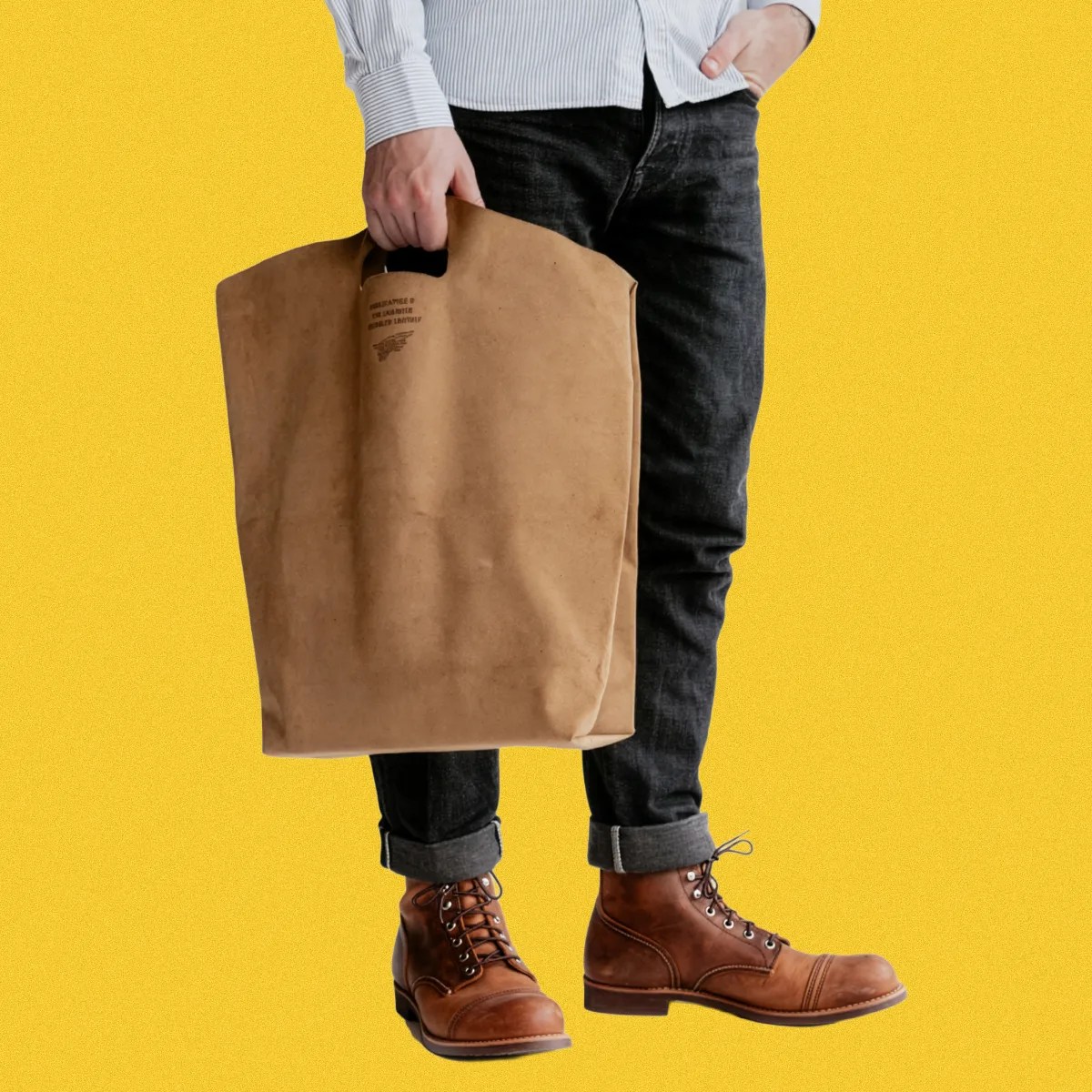 Person holding a large brown leather tote bag with cutout handles, wearing rolled-up black jeans and brown leather boots.
