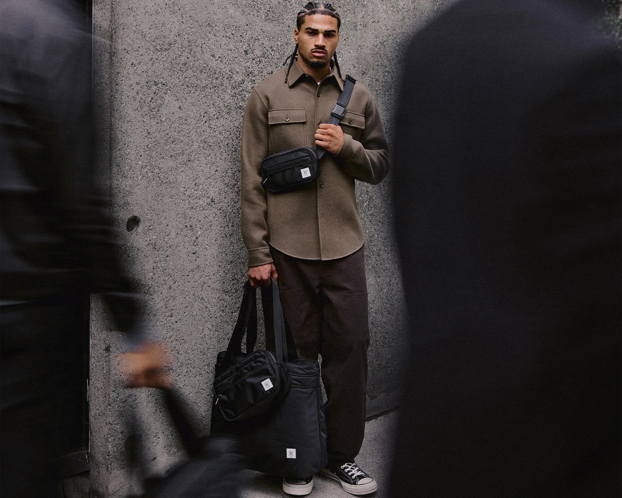Man wearing a brown button-up shirt and dark pants holding a black tote bag and wearing a black crossbody bag.