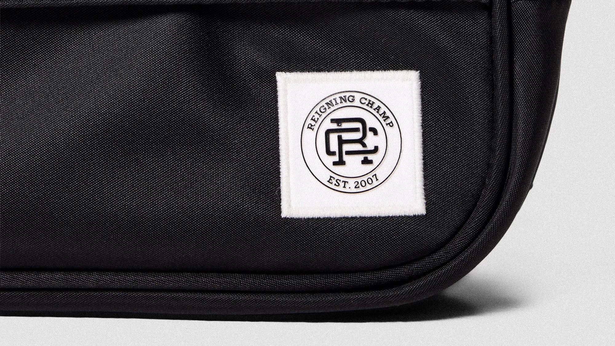 Close-up of a black fabric bag with a white square patch labeled "Reigning Champ EST. 2007" and a circular logo.