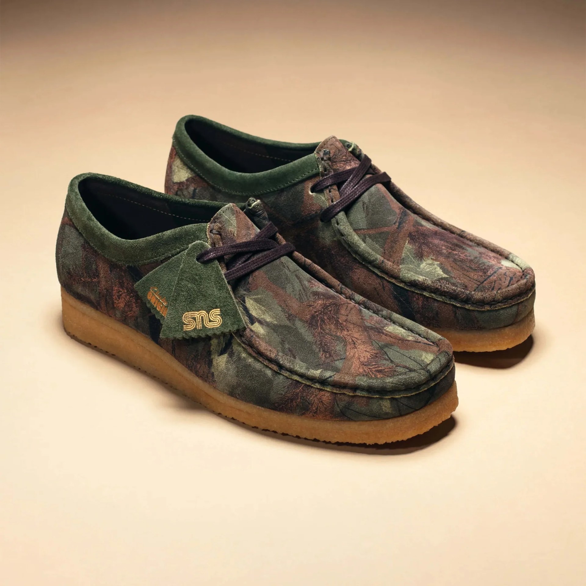 Pair of green and brown camouflage suede moccasin shoes with gum soles and dark laces on a beige surface.