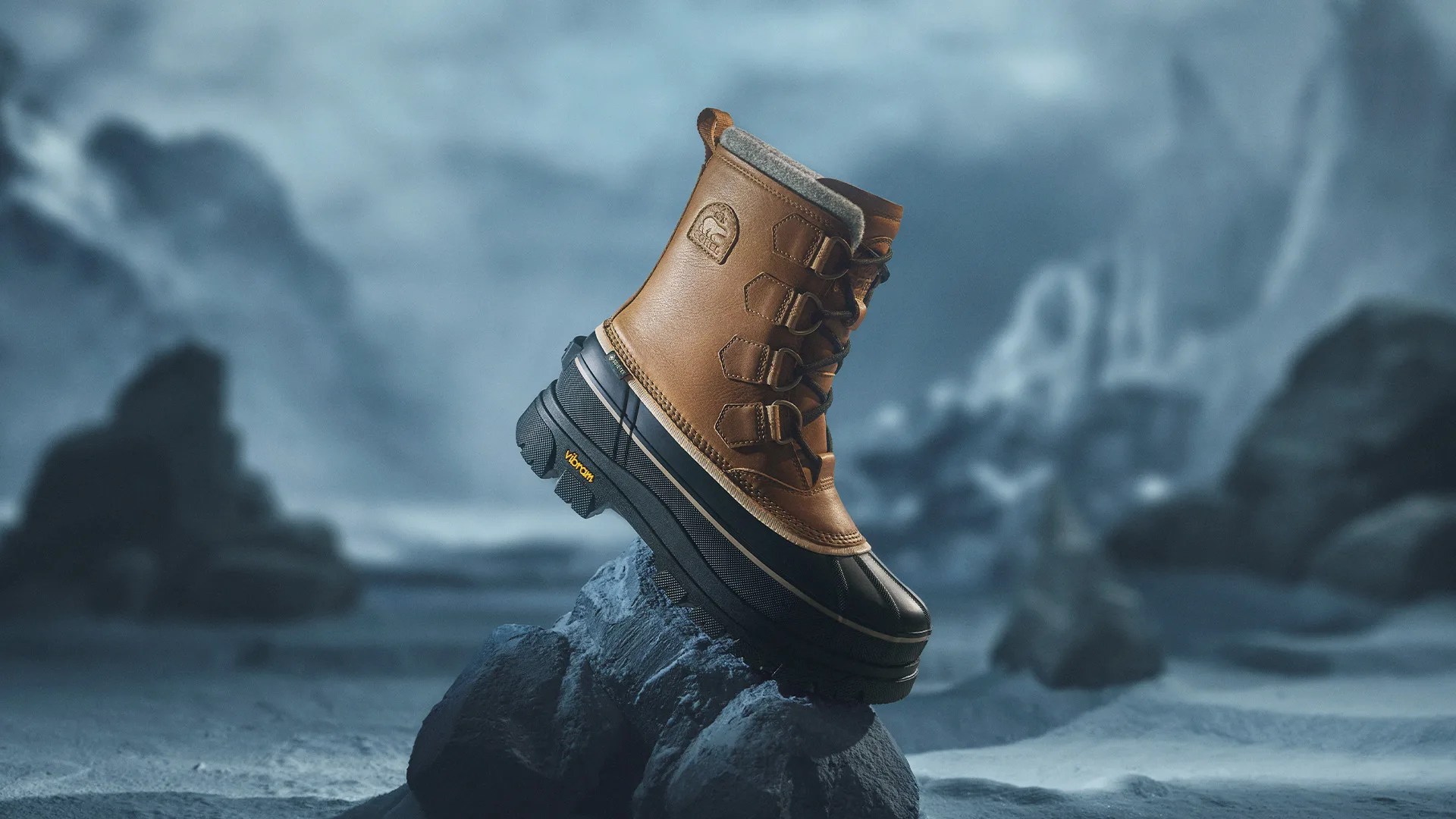 Brown leather and black rubber winter boot with Vibram sole on a rock in a snowy landscape.