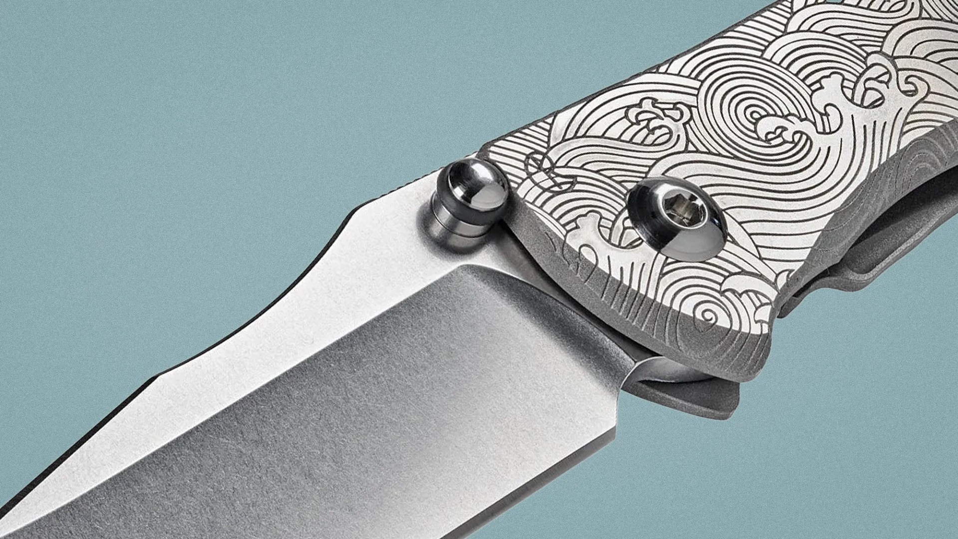 Close-up of a folding knife with a stonewashed blade and a handle engraved with wave patterns.
