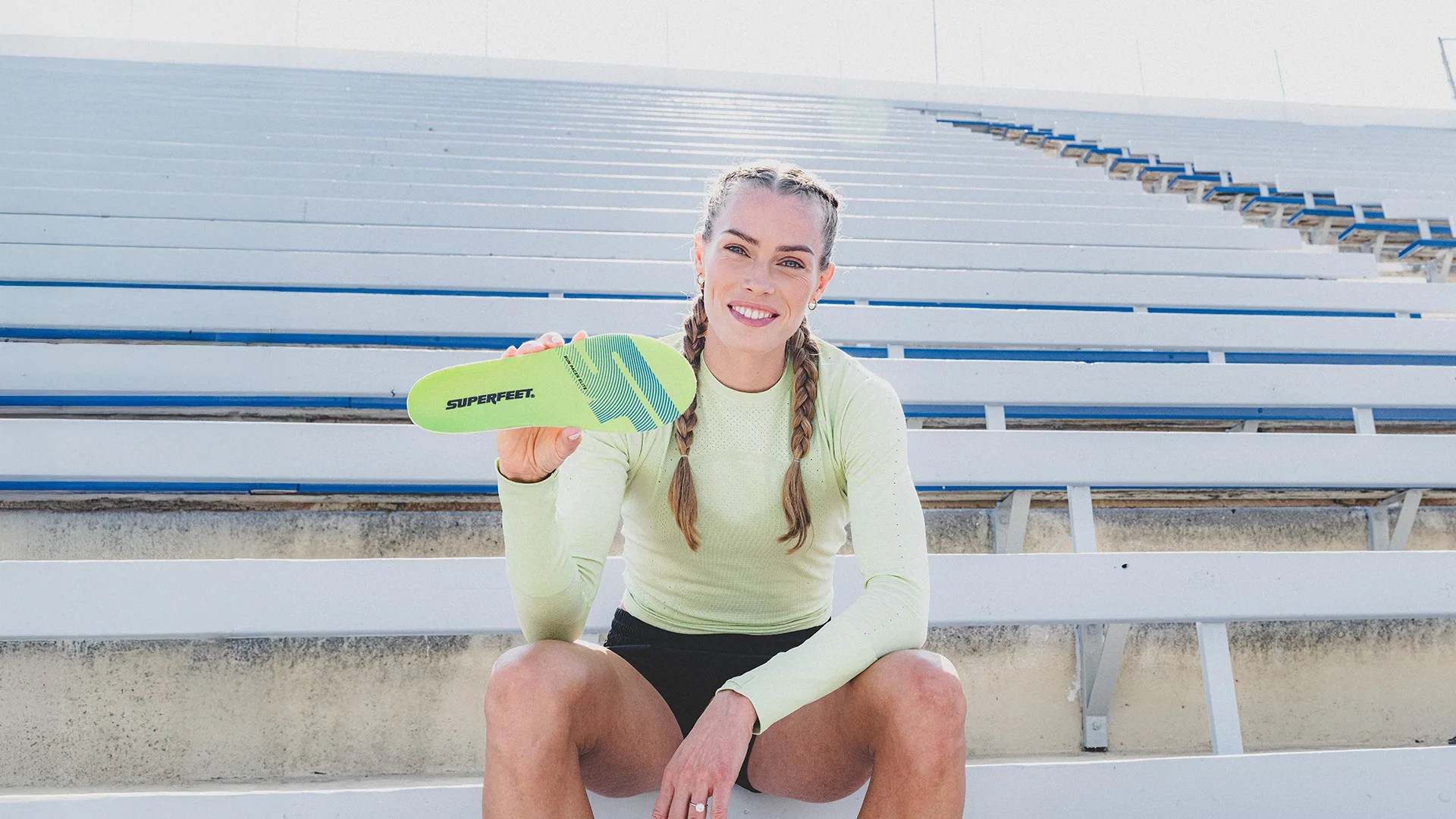 Woman with braided hair sitting on white bleachers holding a lime green Superfeet insole.