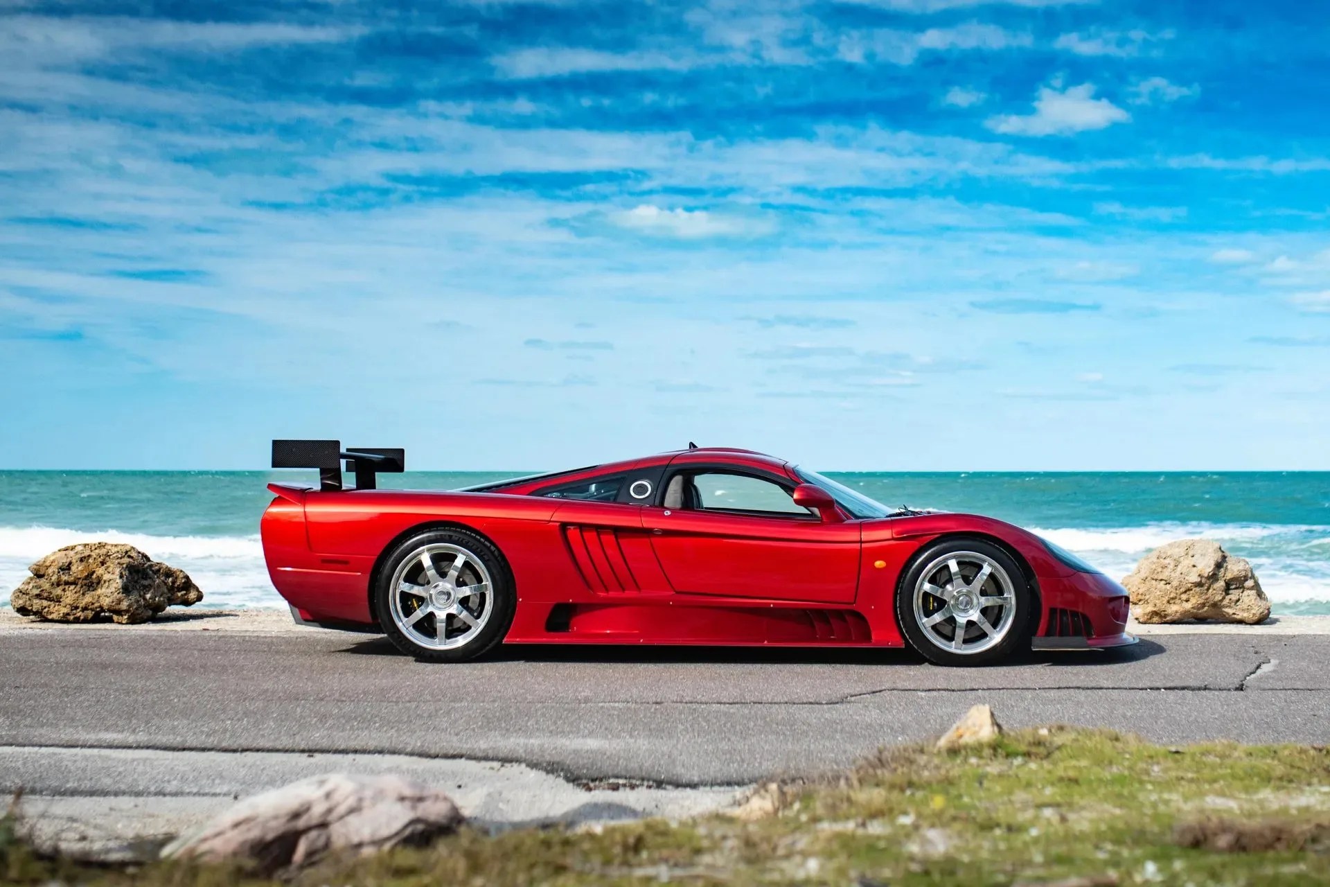 Red low-profile sports car with large rear spoiler parked on a coastal road with ocean and rocks in the background.