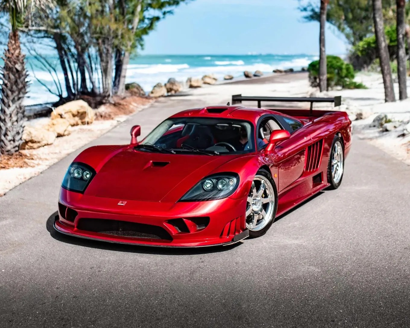 Red sports car with chrome wheels parked on a coastal road with trees and ocean in the background.