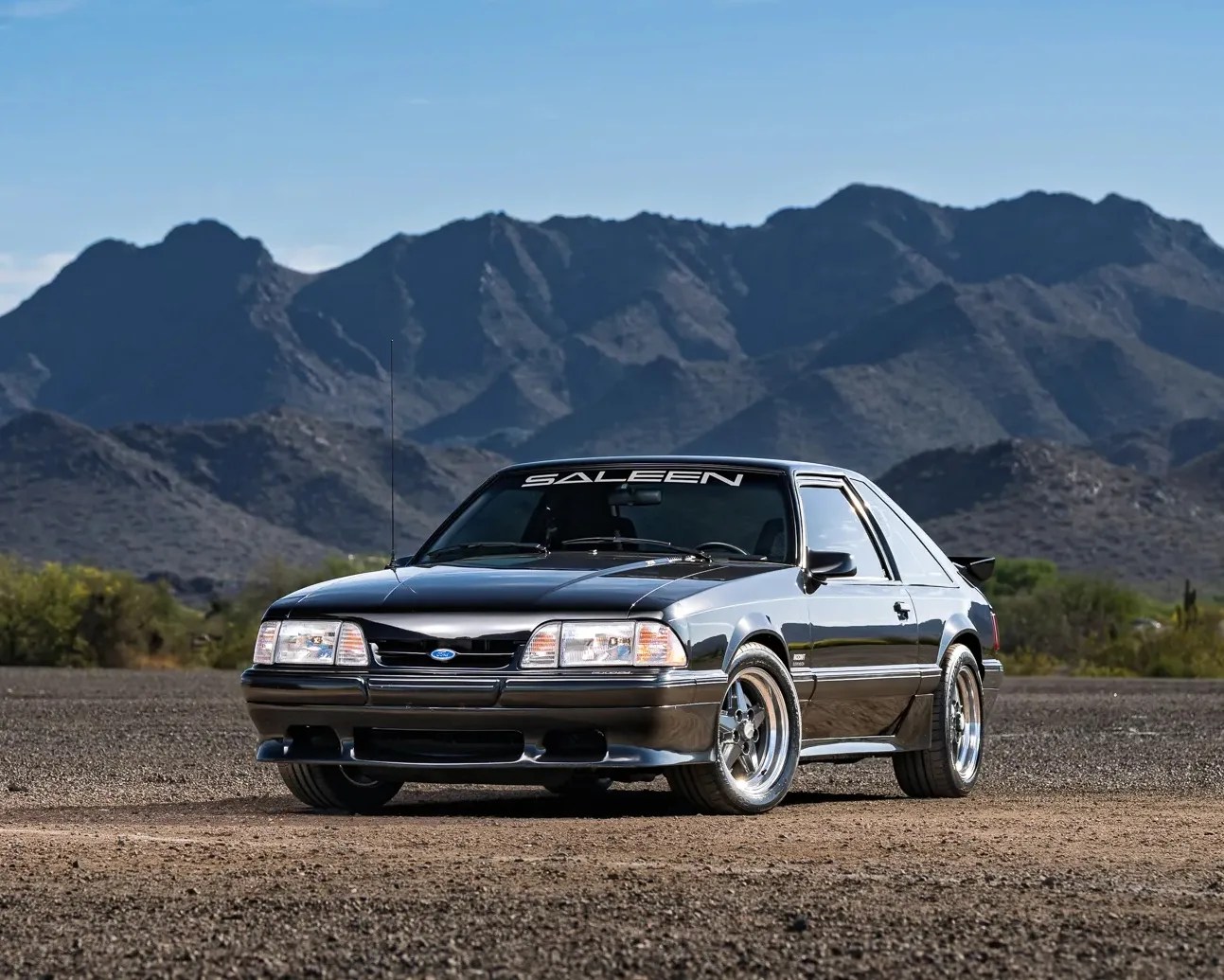 Black and silver Saleen Mustang coupe parked on dirt with mountains in the background.