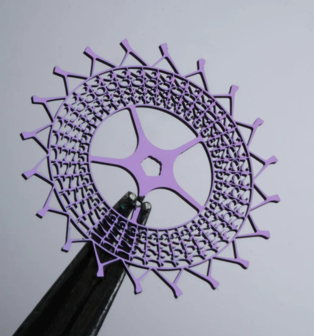 Purple circular gear-like object with intricate cut-out patterns held by black tweezers on a white background.