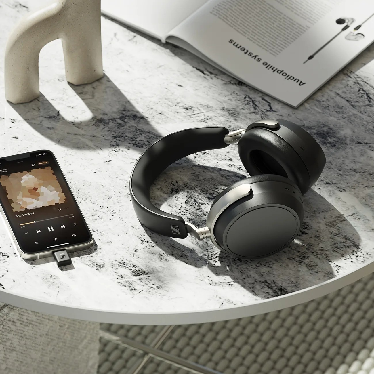Black over-ear wireless headphones with silver accents on a marble table next to a smartphone and an open book.