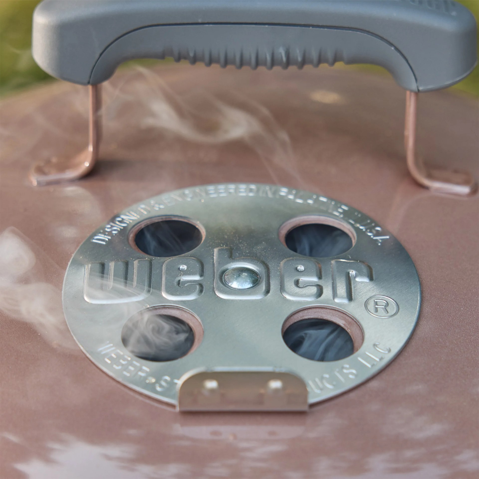 Close-up of a Weber grill lid vent with smoke rising through four circular holes.