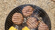 Four grilled hamburger patties and two grilled corn cobs on a round charcoal grill over gravel ground.