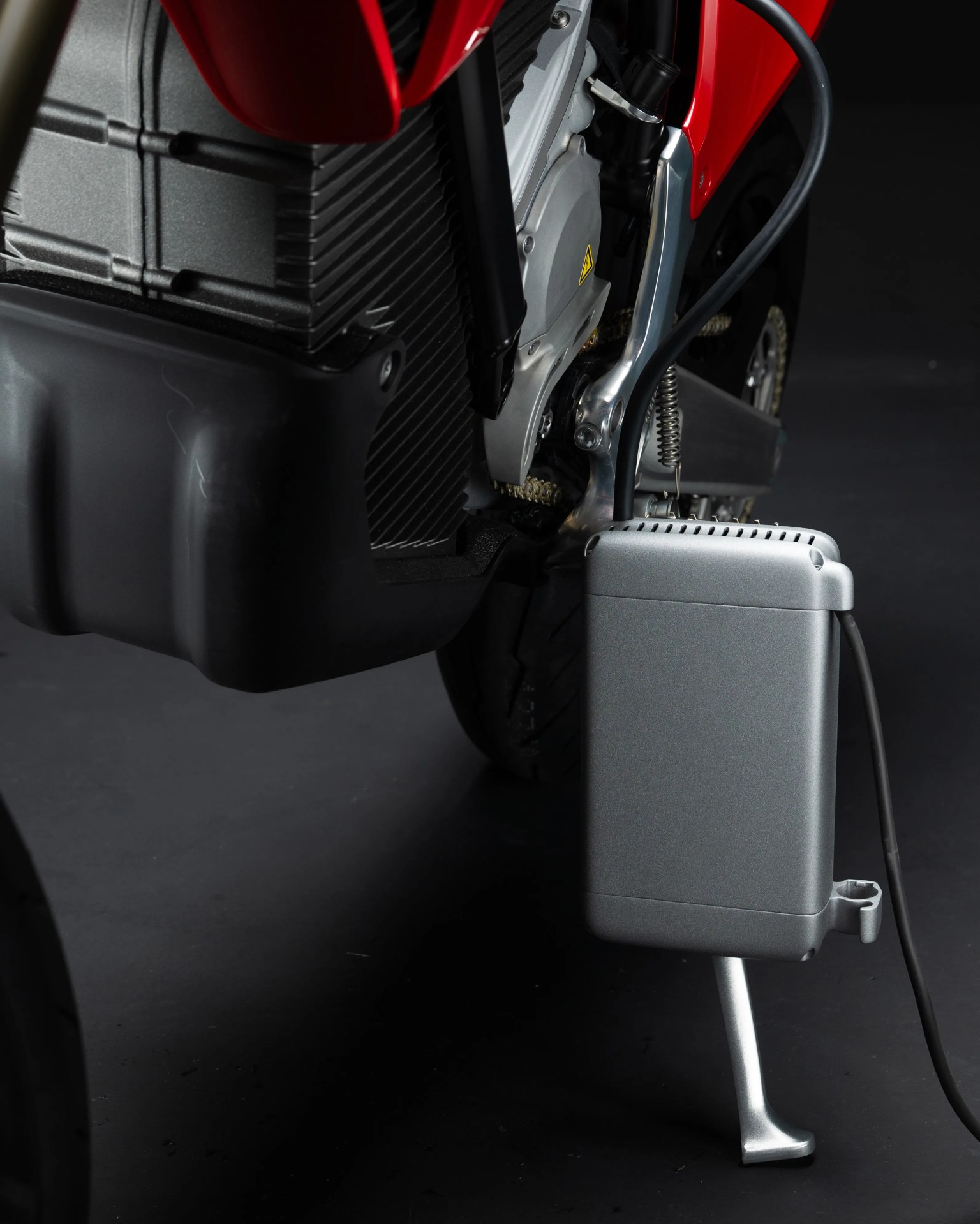 Close-up of a silver rectangular electric motorcycle charger connected near the rear wheel and kickstand.