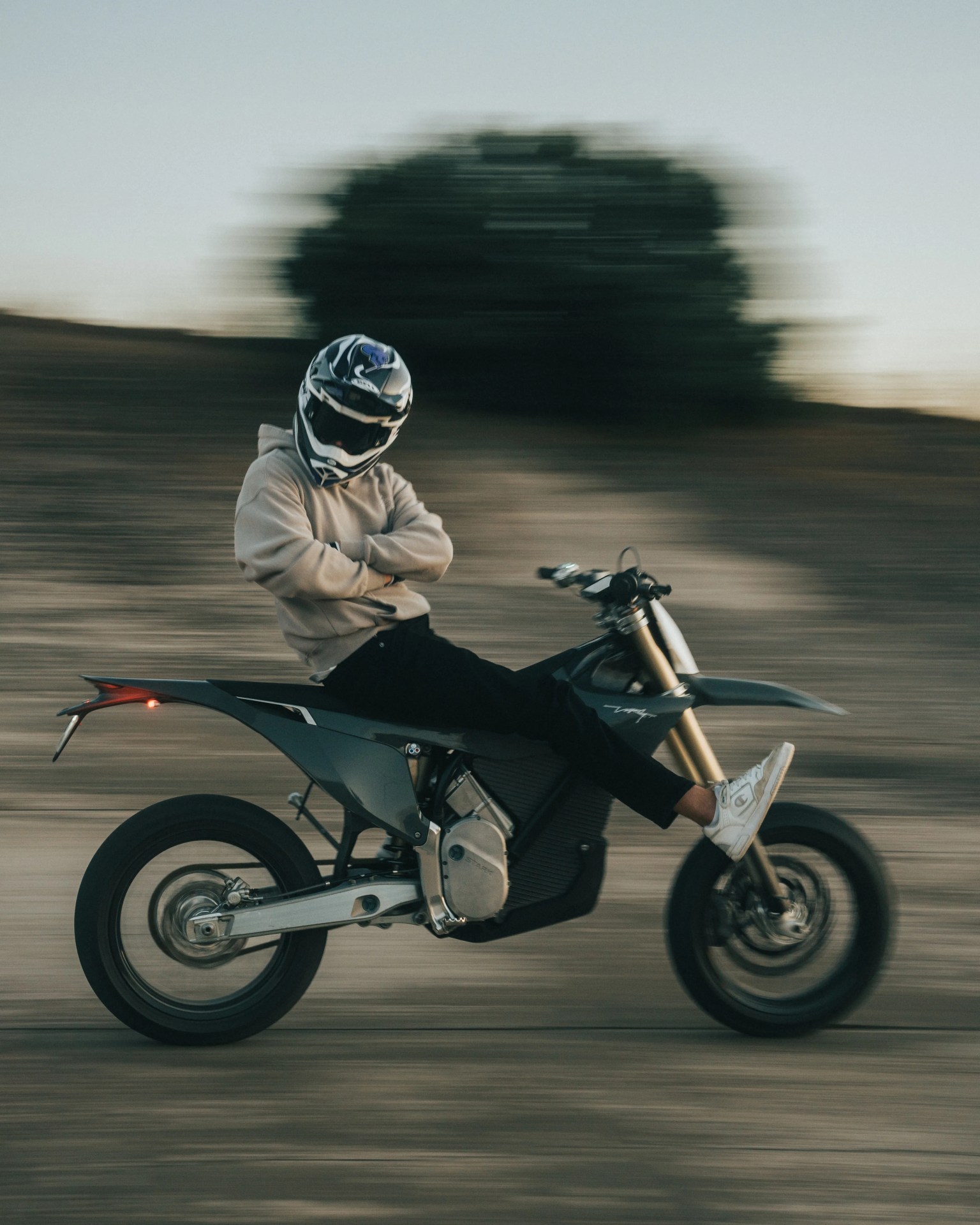 Person wearing a beige hoodie and helmet sitting relaxed with arms crossed on a black dirt bike in motion.