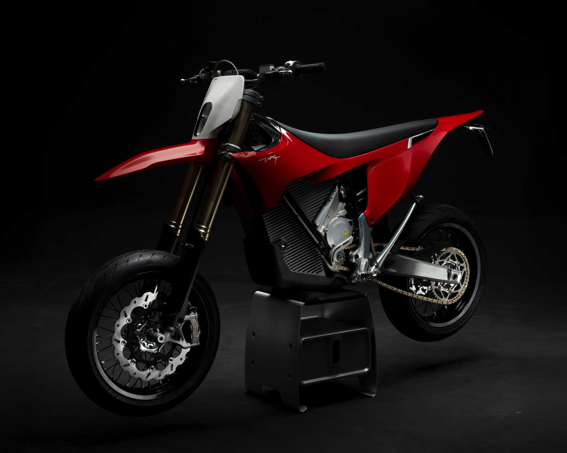 Red and black electric dirt bike with gold front forks and disc brakes on a black stand against a dark background.