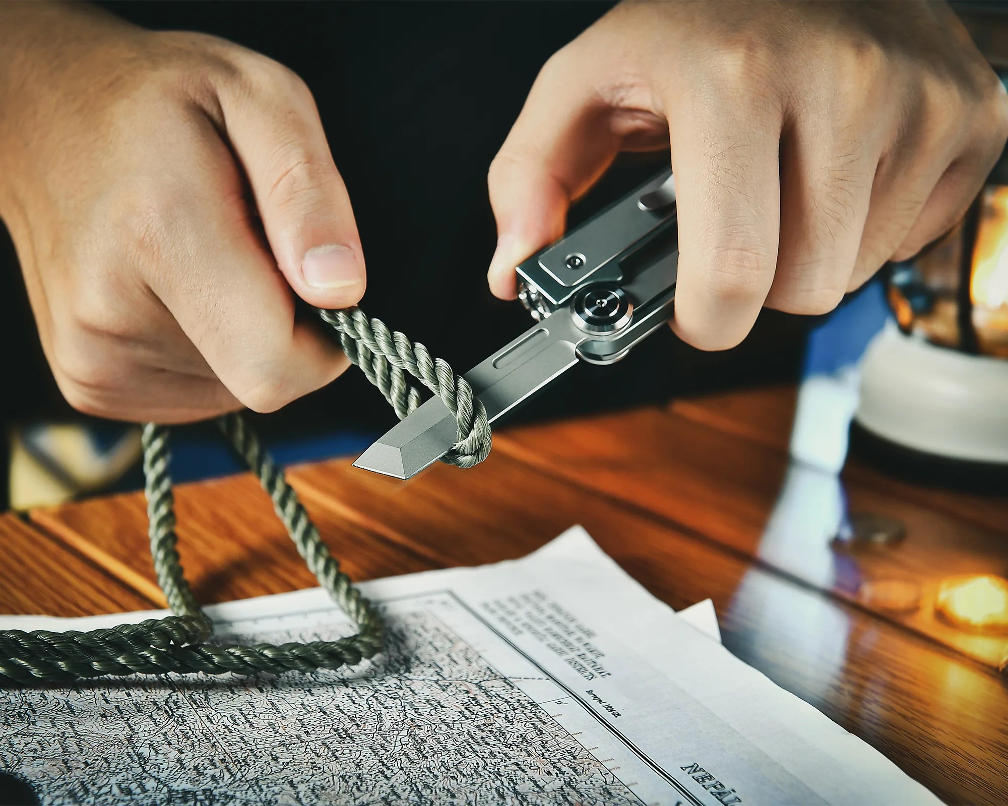Hands holding a silver utility knife cutting a green twisted rope over a map on a wooden table.