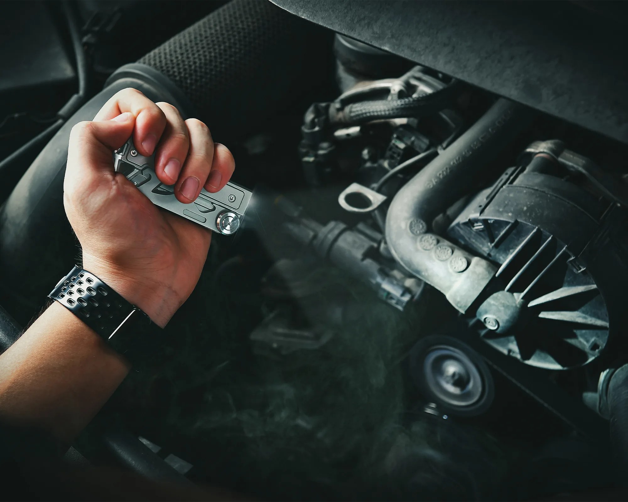 Hand holding a silver flashlight shining light on a car engine with visible belts and hoses.