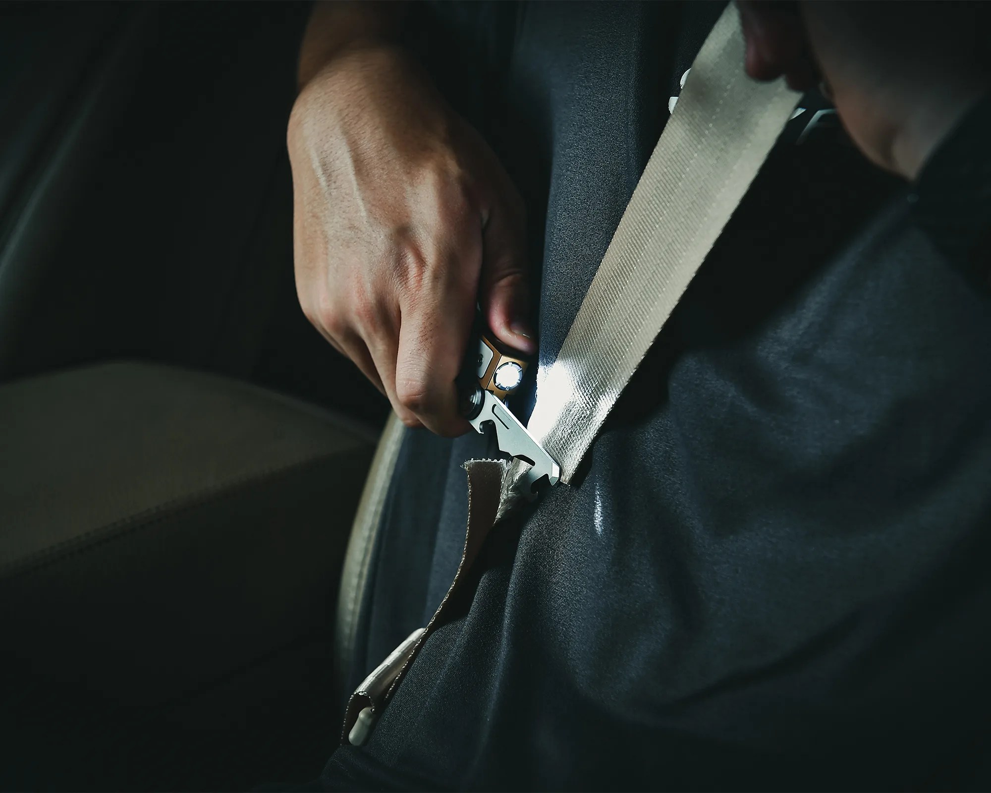 Hand holding a seatbelt cutter tool cutting a beige car seatbelt.