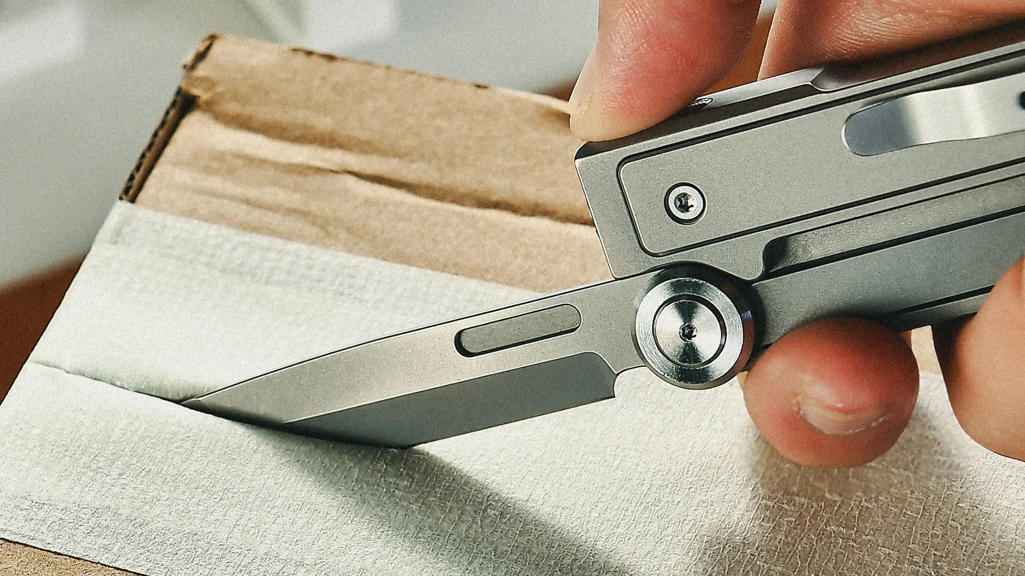 Hand holding a silver folding knife cutting white tape on a cardboard box.
