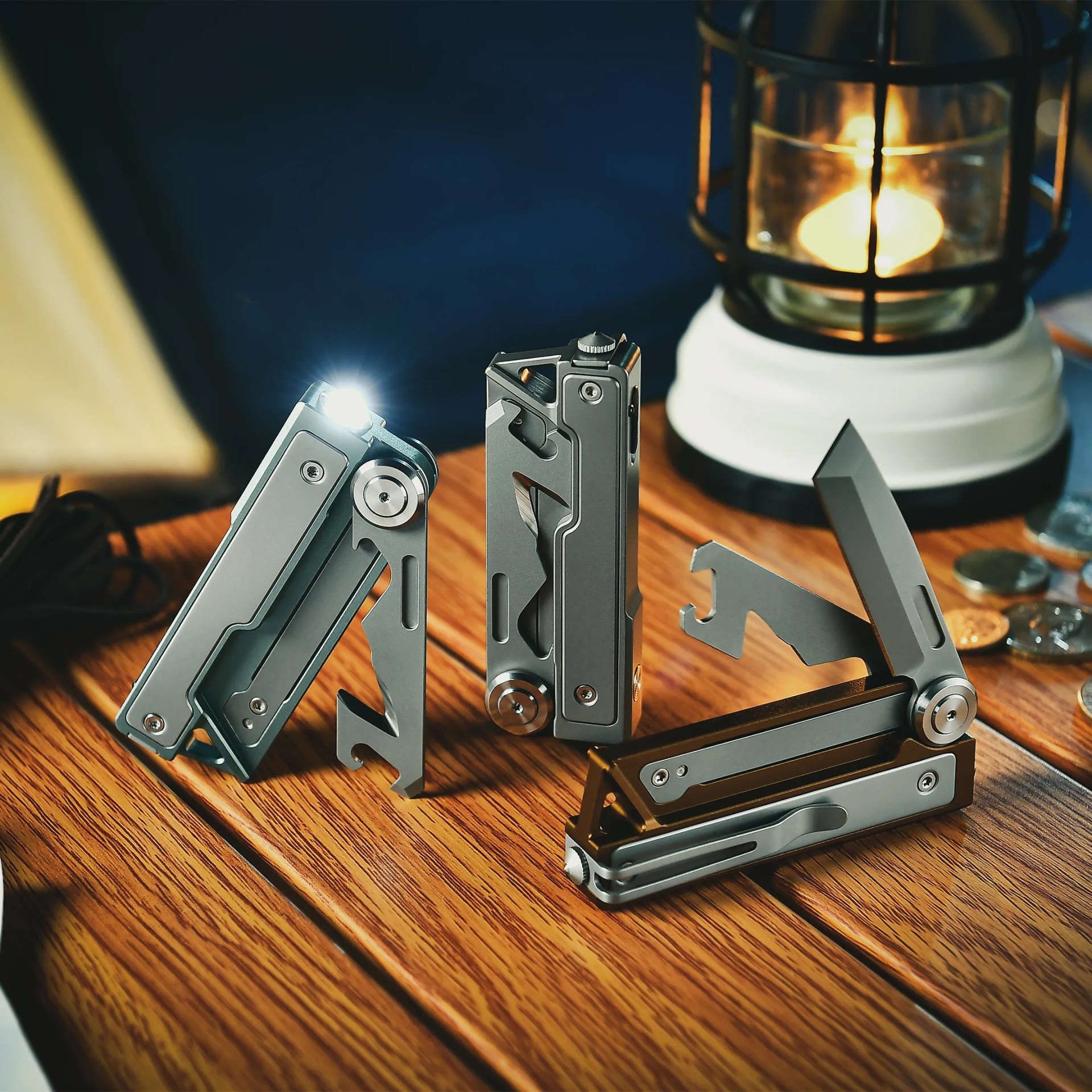 Three gray metal multitools with various tools extended, one with a built-in light turned on, on a wooden surface near a lit lantern.