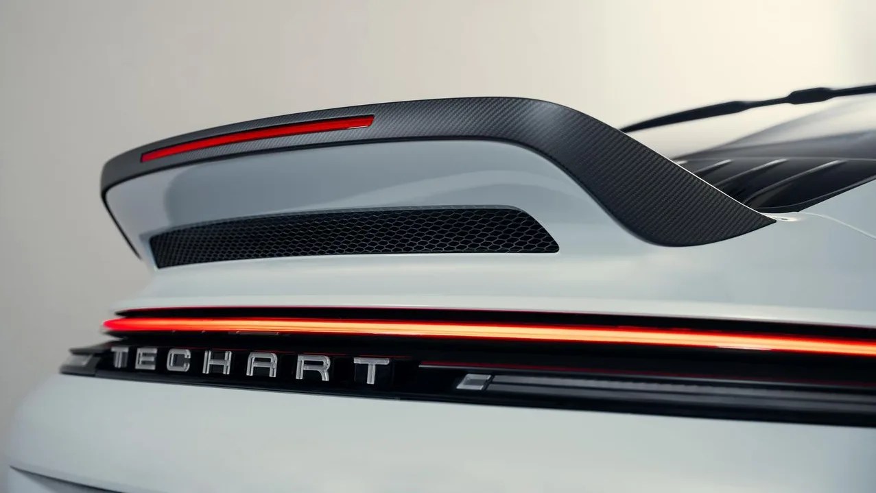 Rear view of a white car with a carbon fiber spoiler and illuminated red tail light strip above the "TECHART" badge.