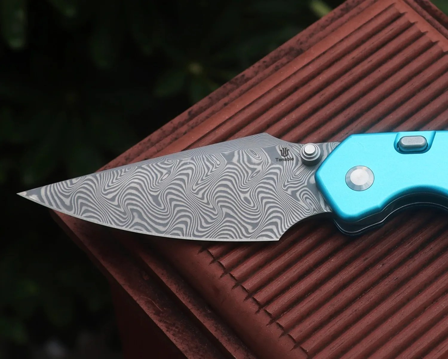 Folding knife with blue handle and patterned Damascus steel blade resting on a textured brown surface.