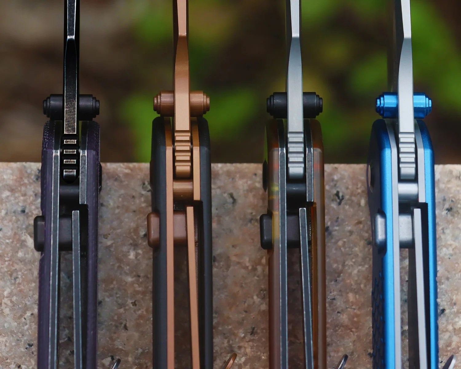 Four folding knives with visible locking mechanisms, in black, bronze, gray, and blue, standing upright on a stone surface.
