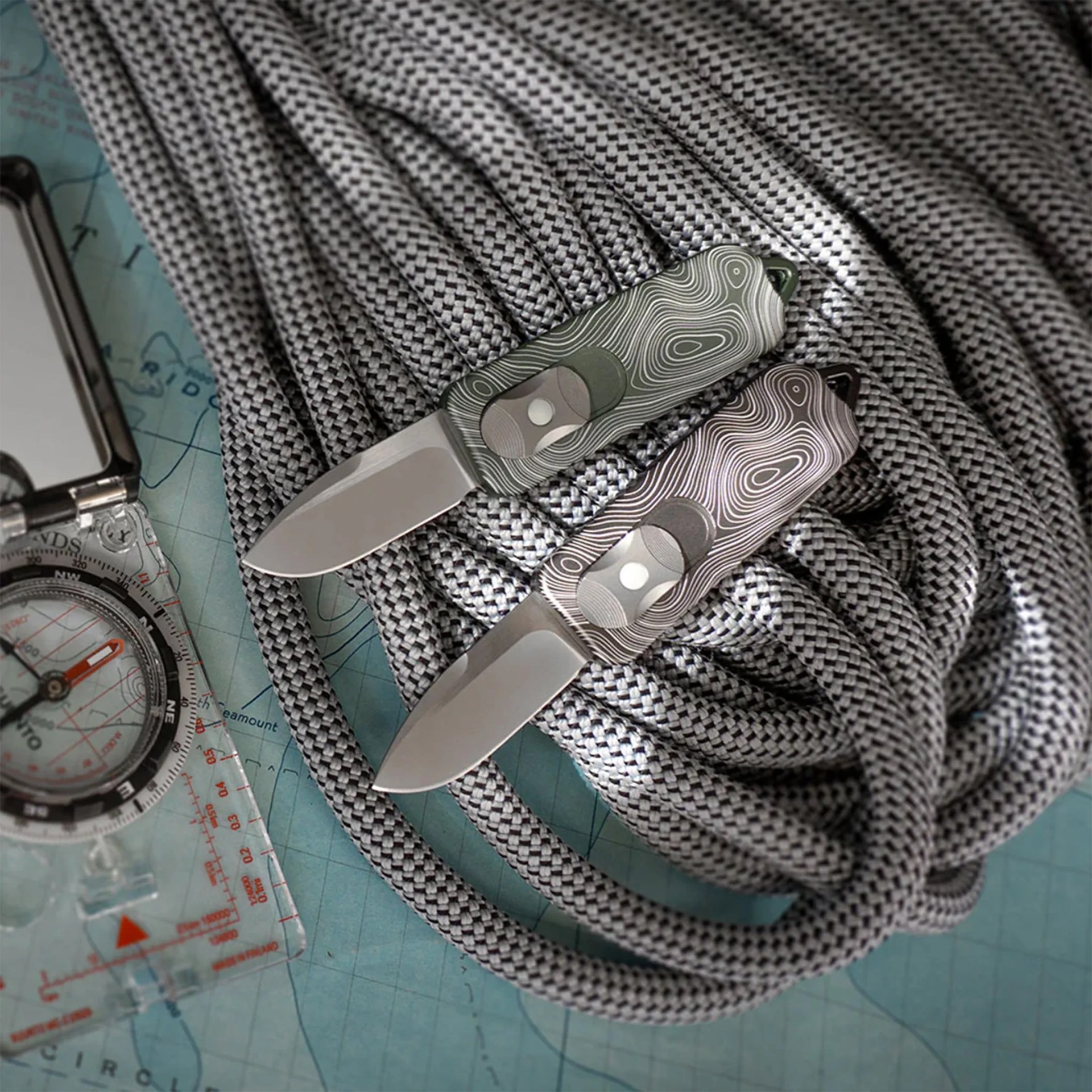 Two small folding knives with patterned handles, one green and one gray, resting on coiled gray climbing rope over a map with a compass.