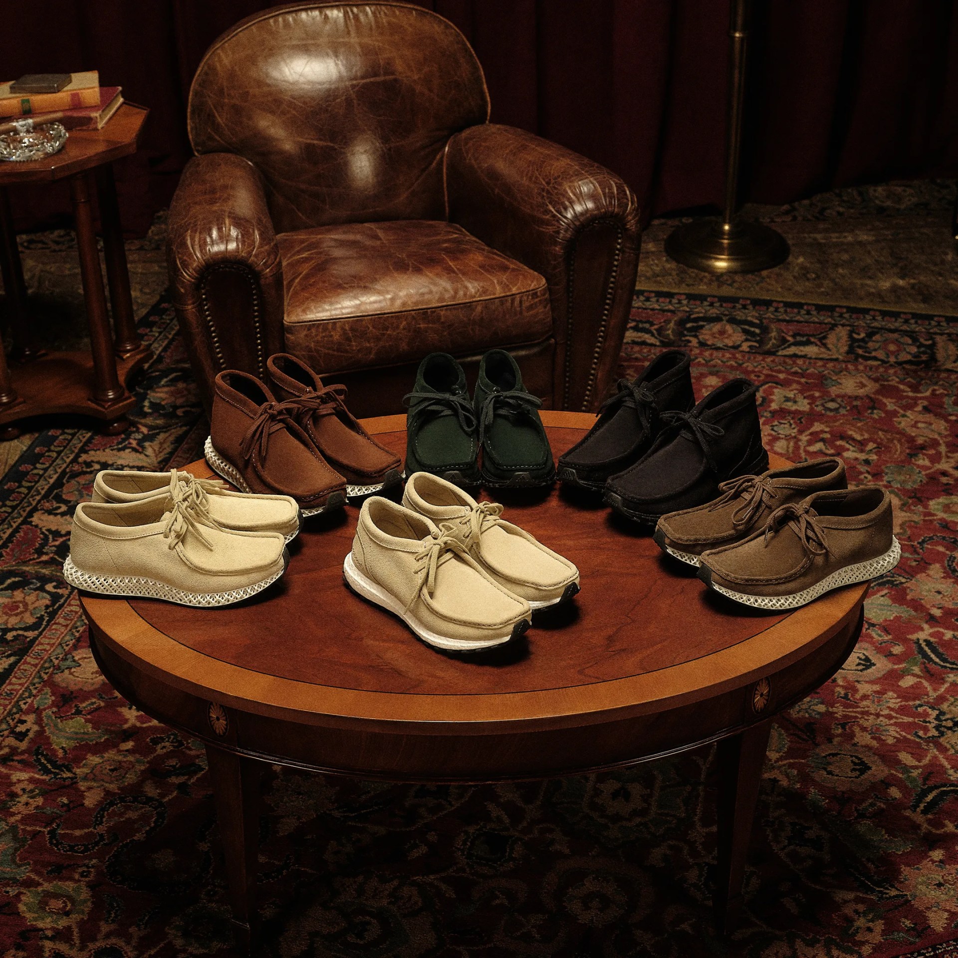 Six pairs of suede lace-up shoes in beige, brown, green, black, and tan arranged on a round wooden table in front of a brown leather armchair.