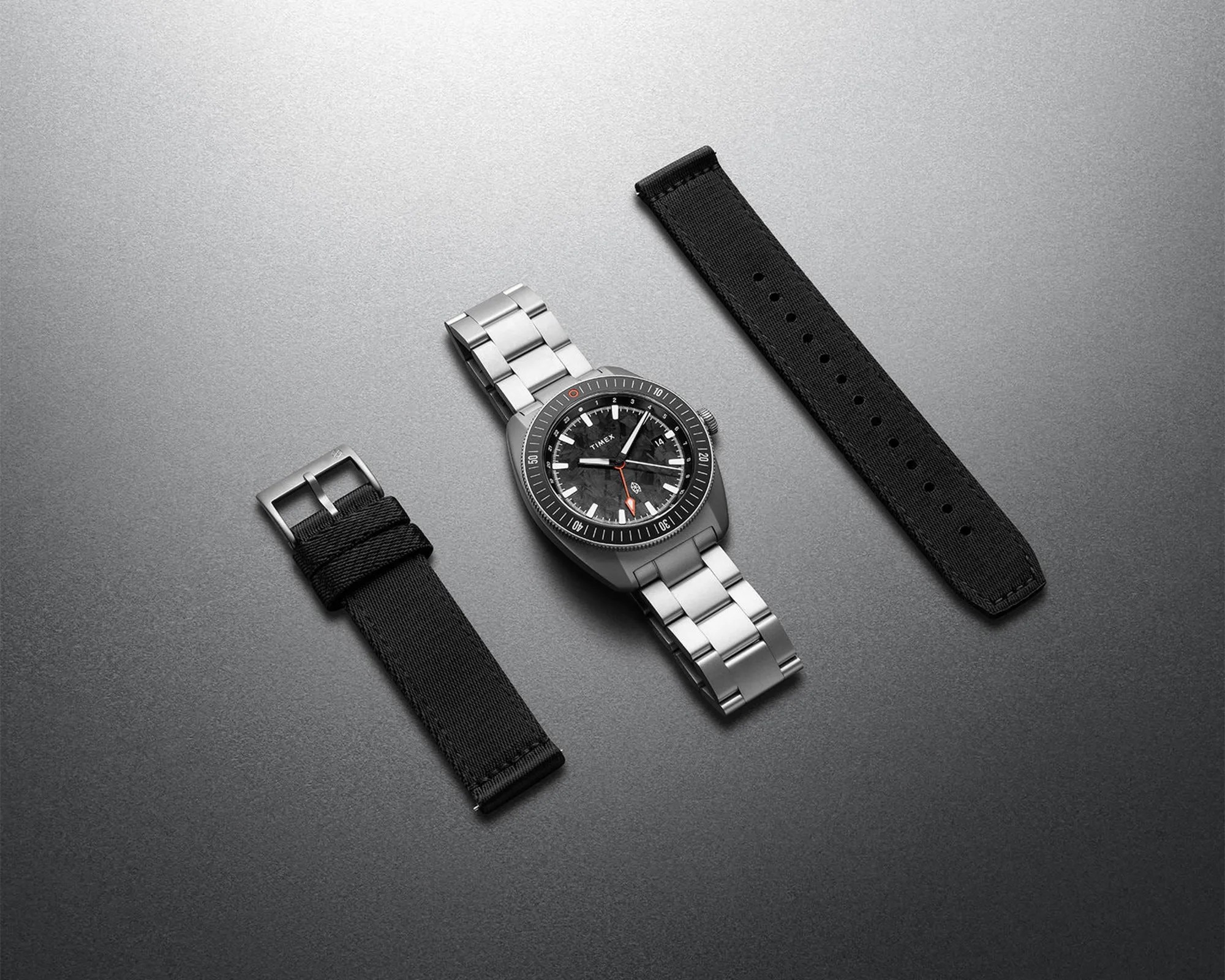 Silver metal Timex wristwatch with black dial and two black fabric replacement straps on a gray surface.