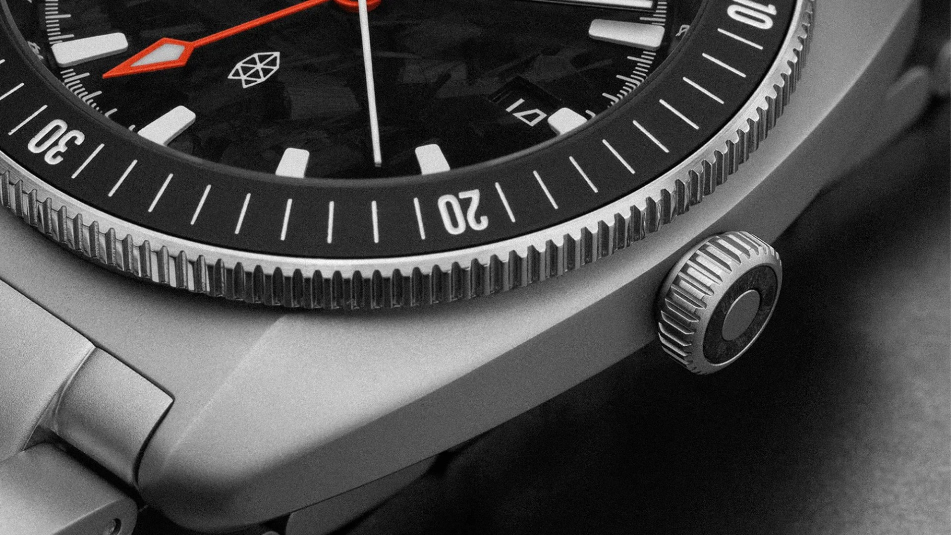 Close-up of a silver metal watch bezel and crown with a black dial featuring white markers and an orange hour hand.
