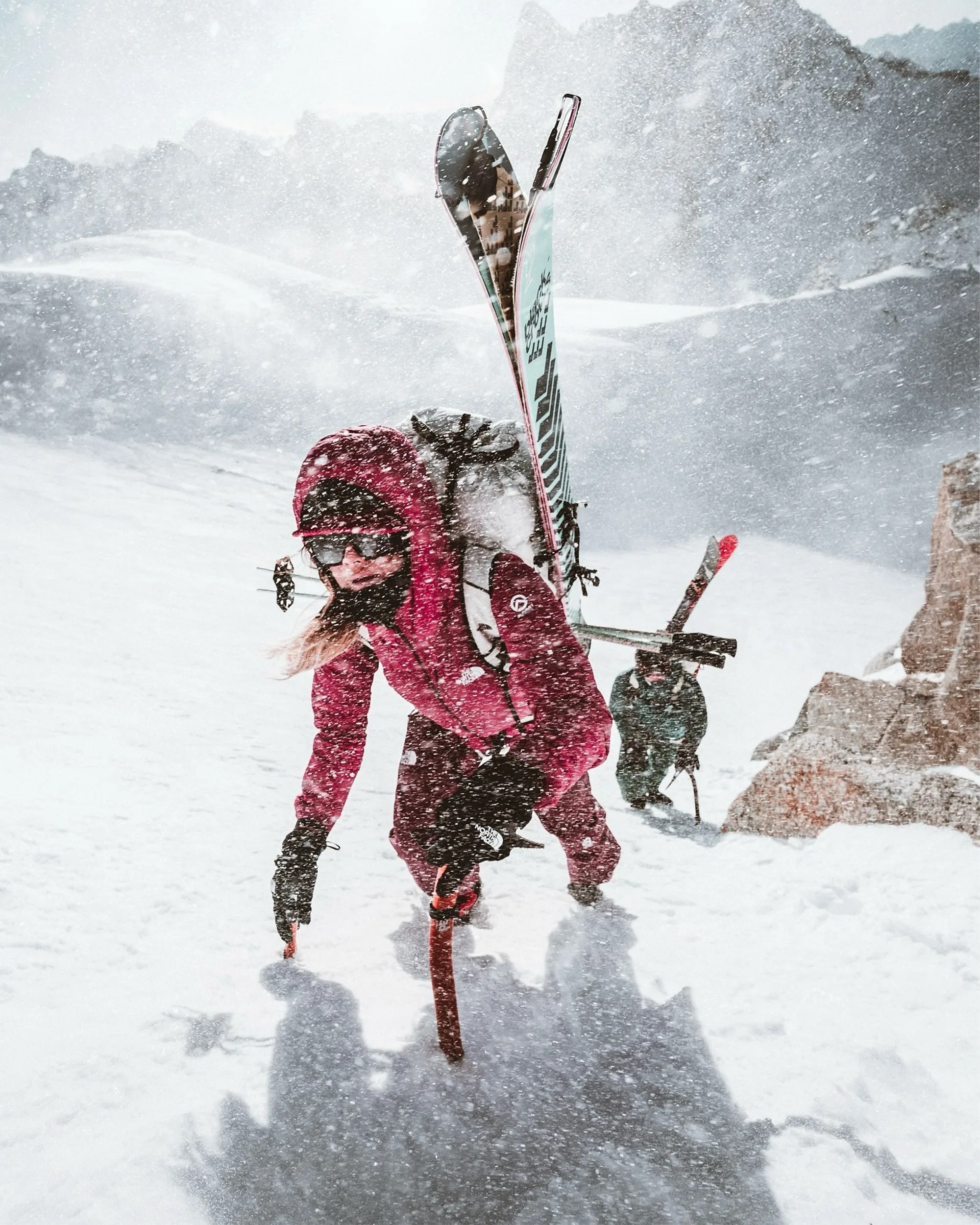 Person in red winter gear climbing snowy mountain with skis on backpack during snowstorm.