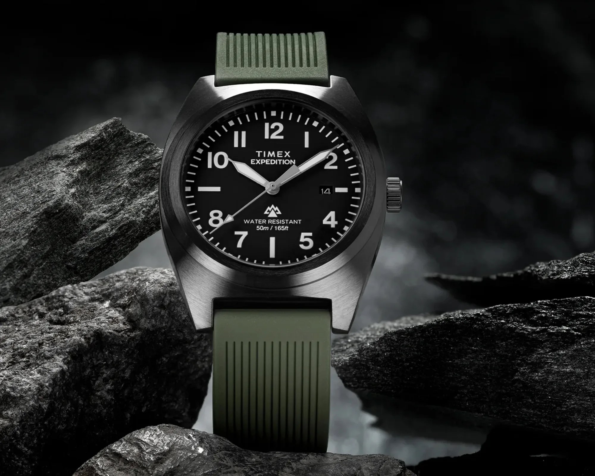 Timex Expedition watch with black dial, silver case, and green ribbed silicone strap on dark rocks.