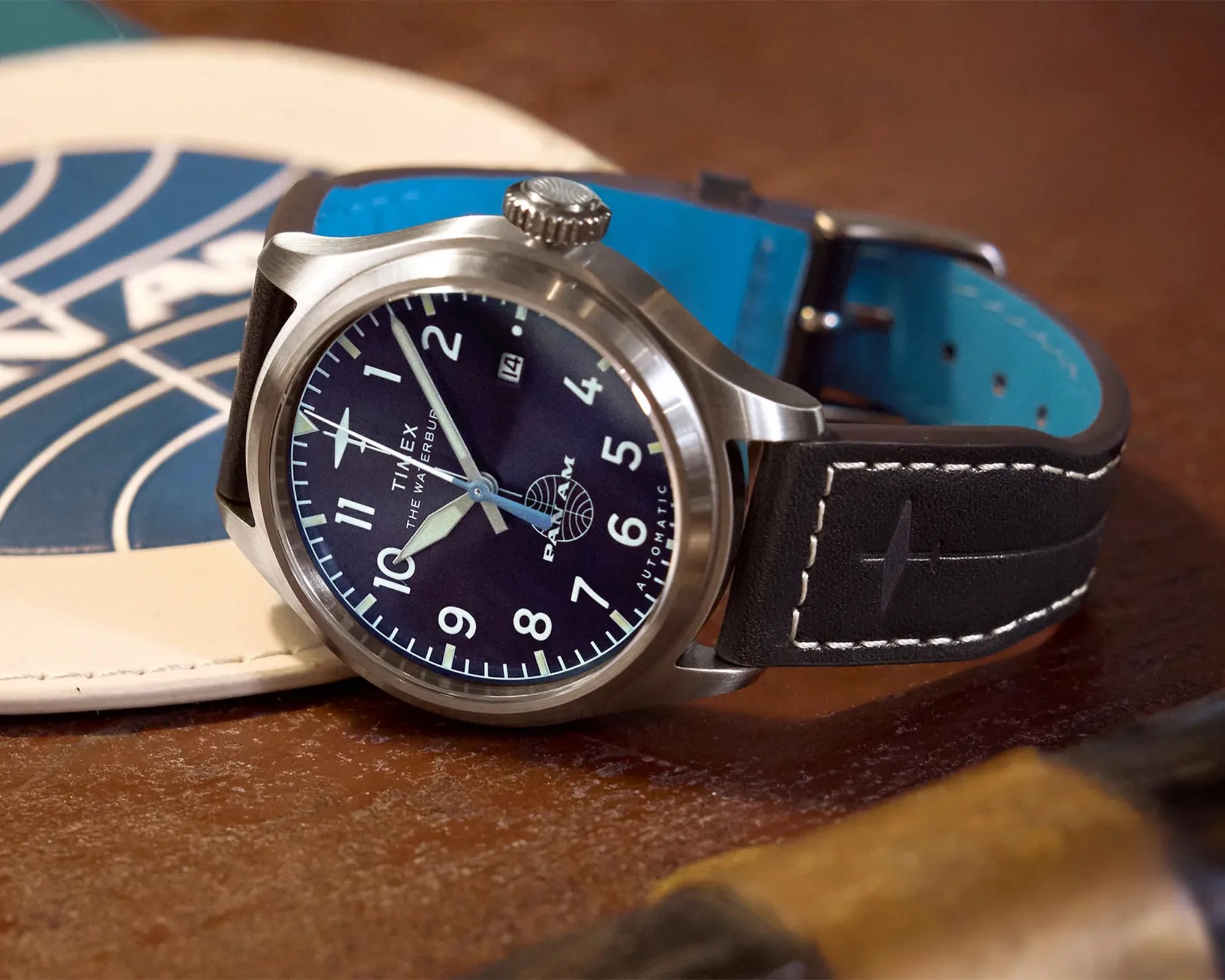 Timex wristwatch with black dial, white numerals, date window, and black leather strap with blue lining on a brown surface.