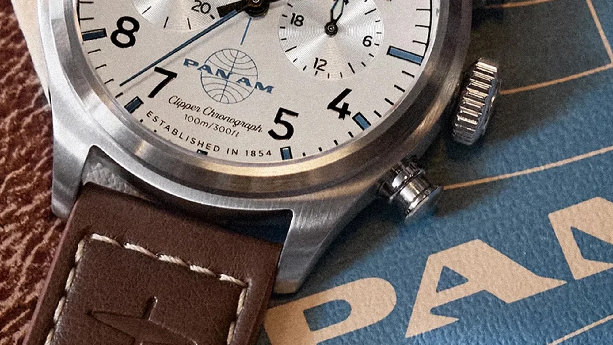 Close-up of a Pan Am Clipper Chronograph watch with a white dial, black numerals, blue accents, and a brown leather strap with white stitching.