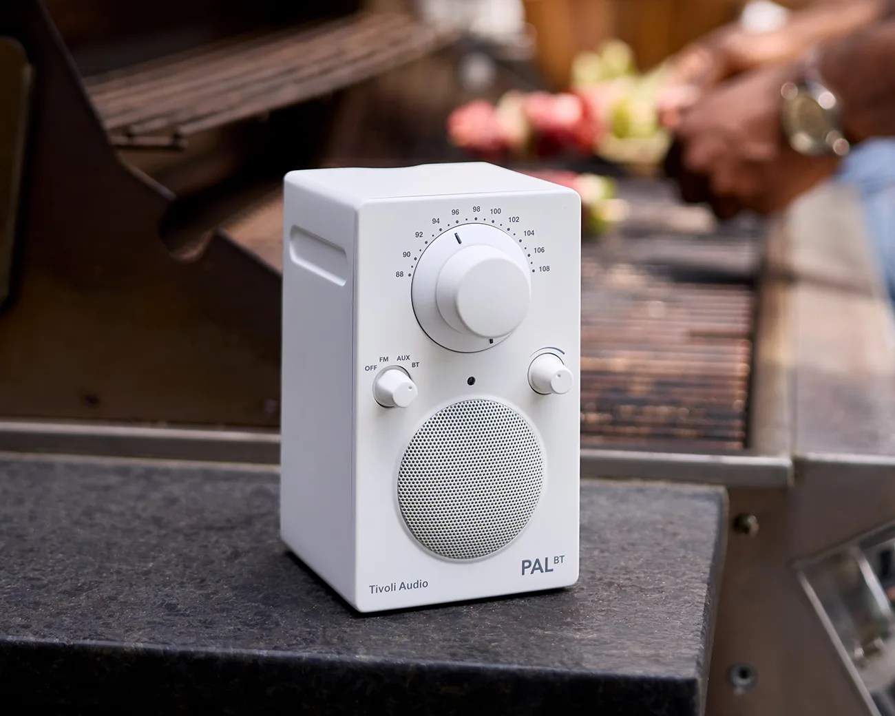 White Tivoli Audio PAL BT radio with dials and speaker on a dark surface near a grill.